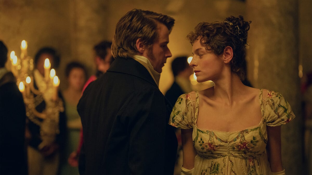 Jack Lowden, seen from behind in a dark coat, looks at Emma Corrin, in a high-waisted floral dress, in a candlelit ballroom.