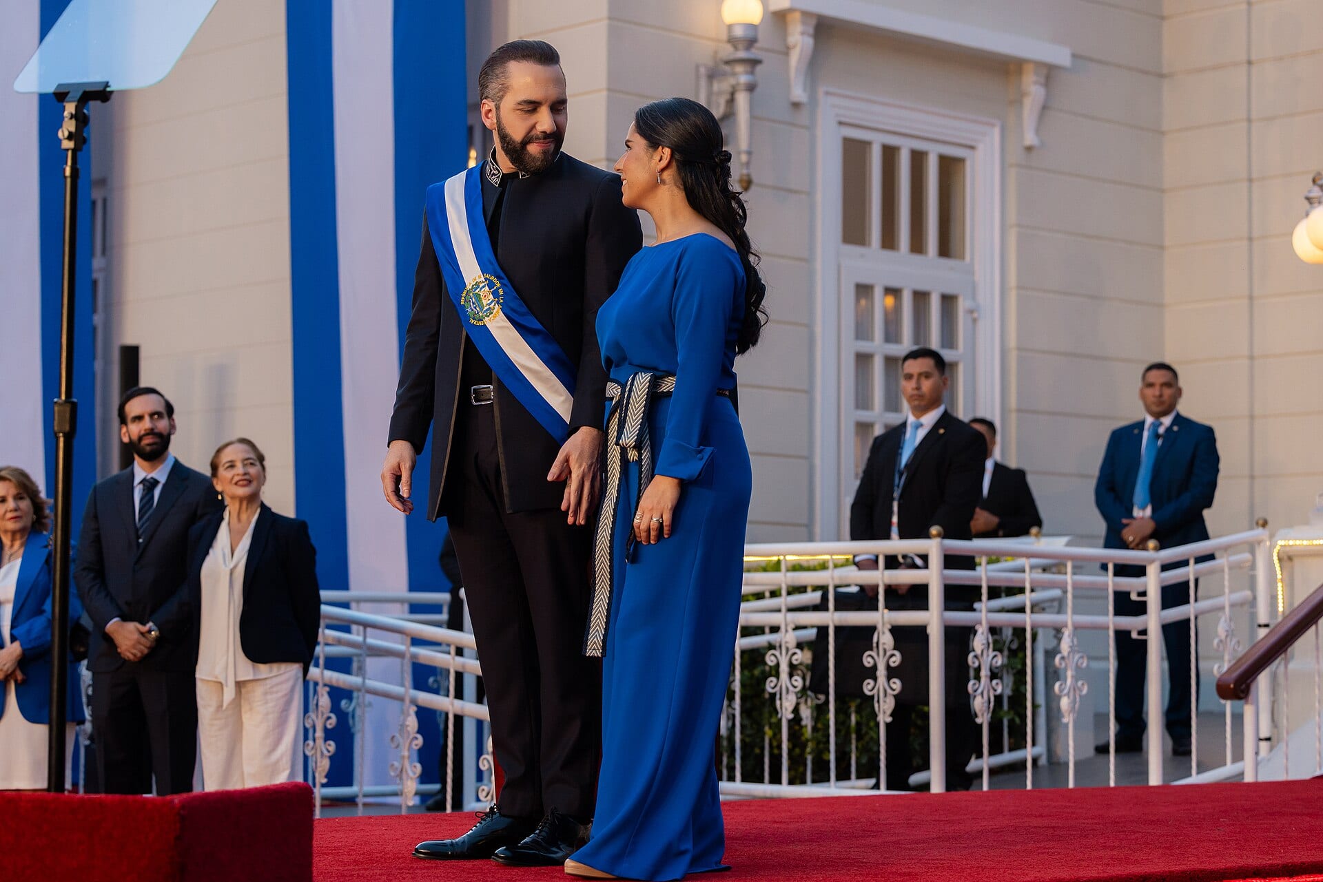 Nayib Bukele, con la banda presidencial azul y blanca, mira a una mujer de vestido azul sobre una alfombra roja frente a un edificio blanco.