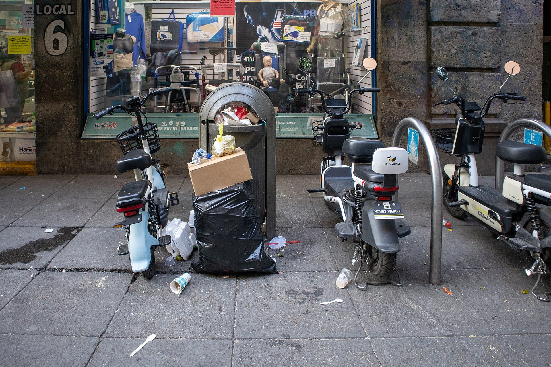 Dos motonetas eléctricas estacionadas a los lados de un bote de basura lleno, frente a un local de una calle del Centro.