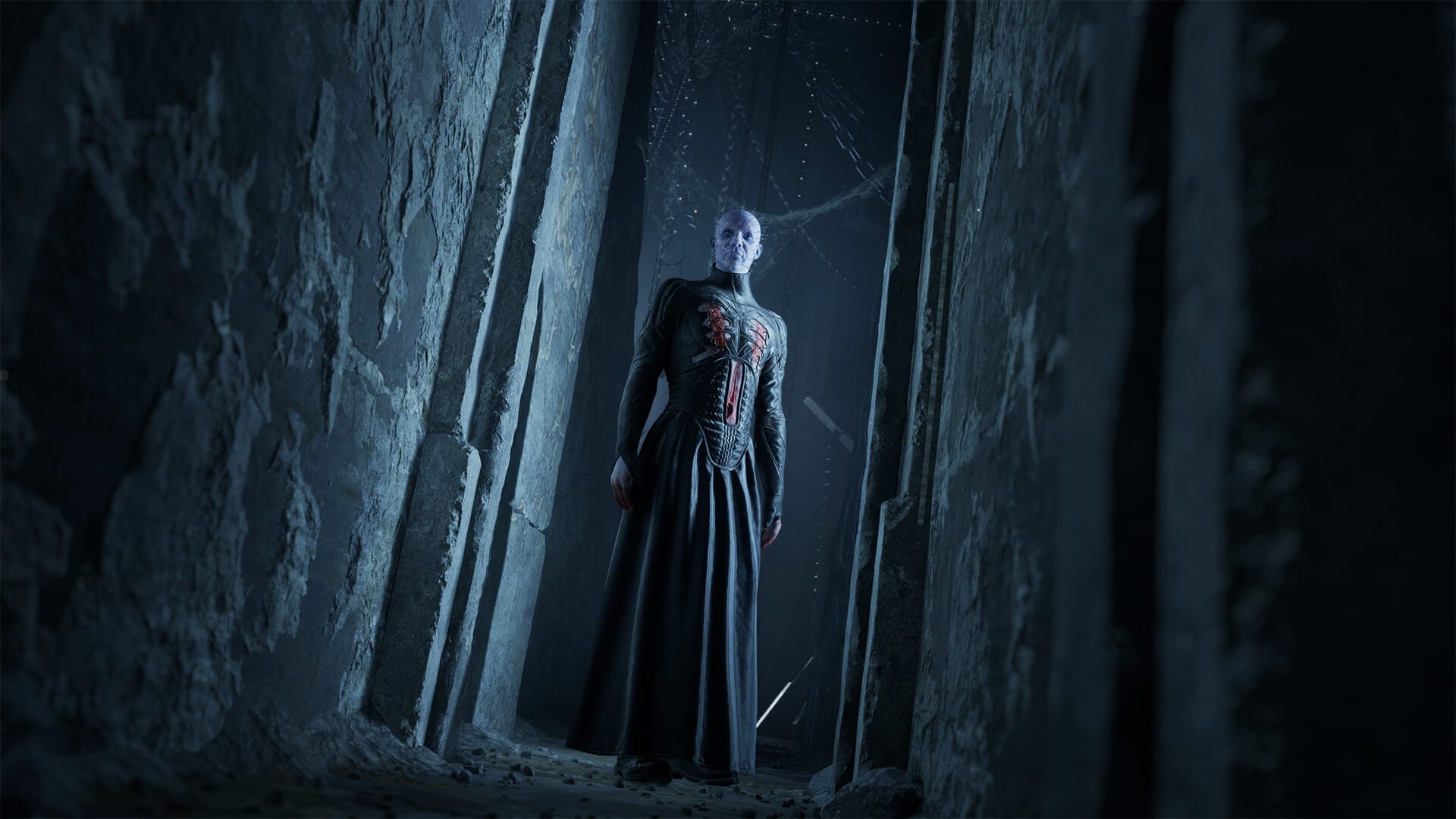 Pinhead, pale-skinned and wearing a long black gown with red stitching on the chest, stands at the end of a narrow rock corridor lit in blue.