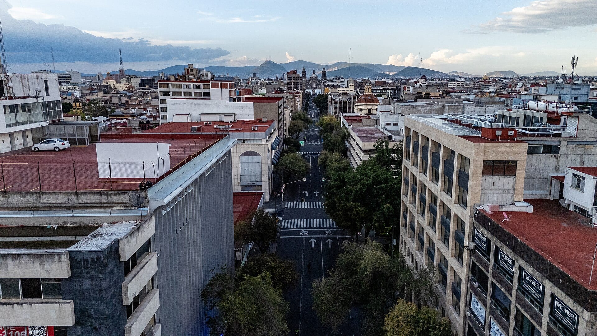 Vista aérea de una avenida con árboles entre edificios del Centro de la Ciudad de México, con cerros al fondo y un cielo con nubes.