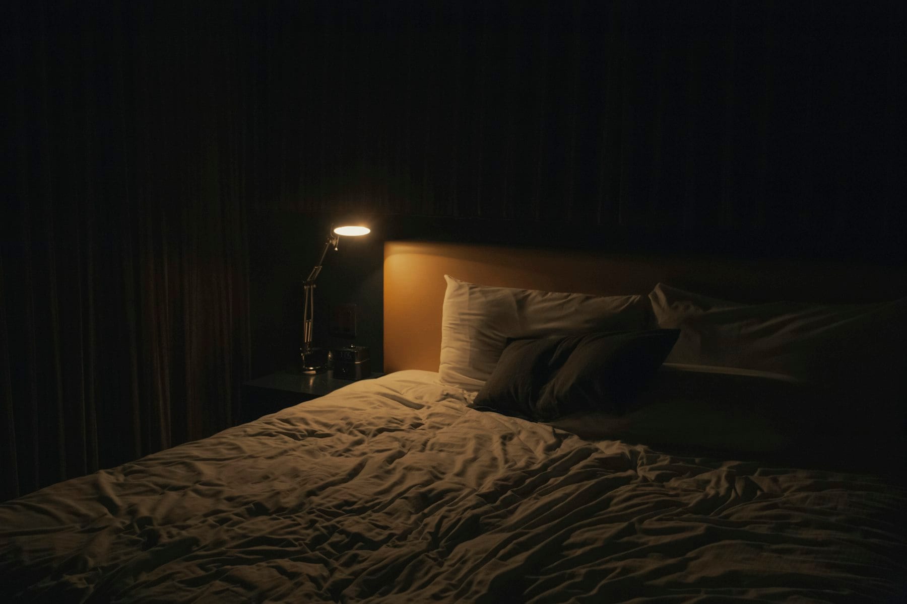 Cama con sábanas blancas arrugadas y almohadas en una habitación a oscuras, iluminada solo por una lámpara de escritorio encendida sobre la mesa de noche.