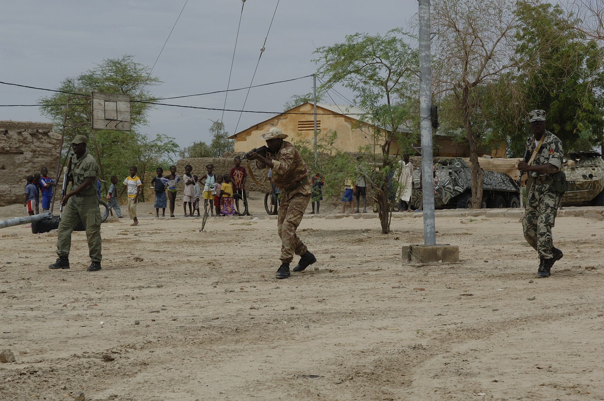 Three soldiers with rifles advance across a dirt square as a group of children watch from behind and two armored vehicles wait beside some trees.