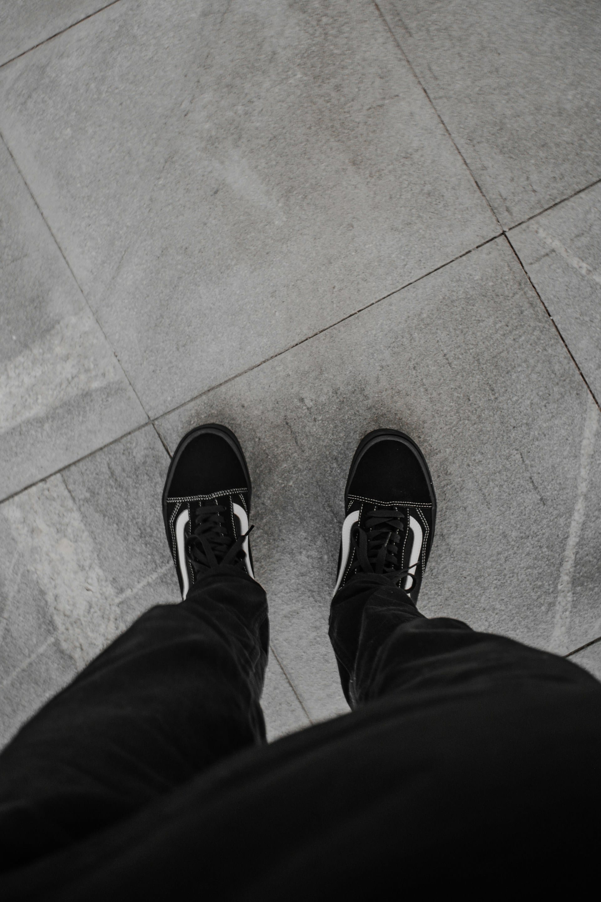 Vista desde arriba de dos pies con zapatillas negras sobre un piso de losas grises.