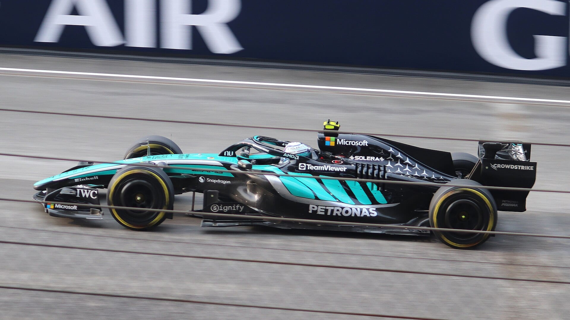 A green and black Mercedes F1 car speeds past a blue advertising board.