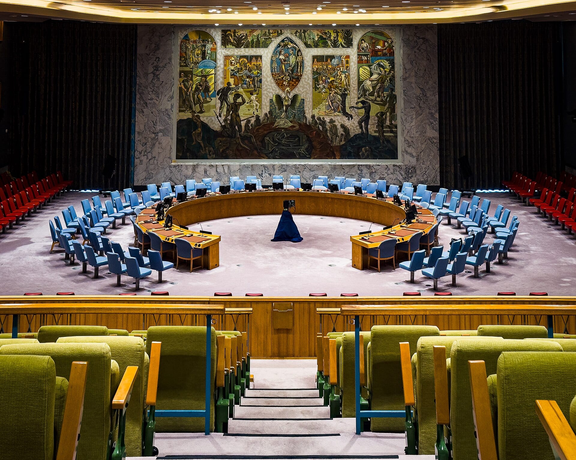 Sala vacía del Consejo de Seguridad de la ONU, con la mesa semicircular, sillas azules y un mural al fondo.