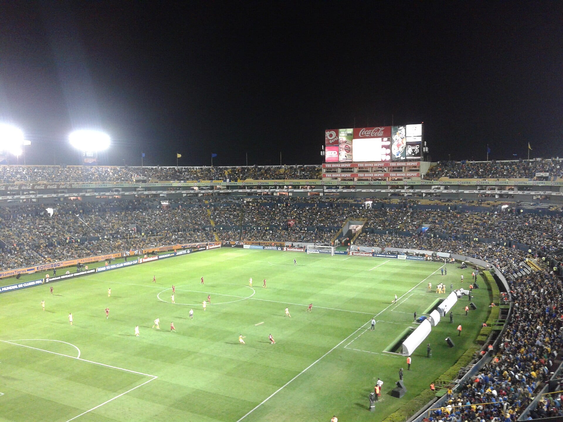 Vista nocturna del Estadio Universitario con las tribunas llenas, un marcador al fondo y jugadores sobre el césped.