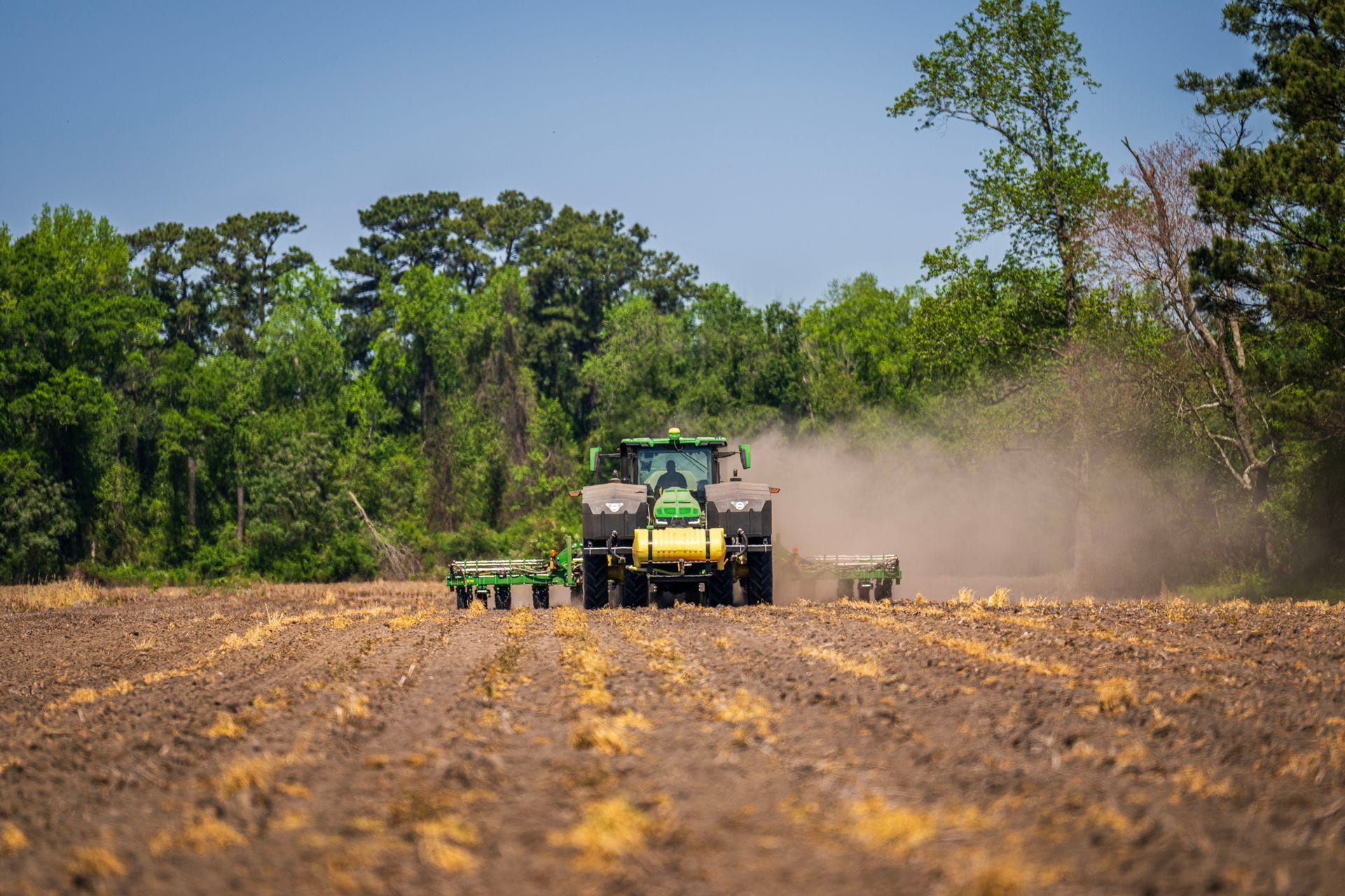 Un tractor verde con implementos avanza por un campo arado y levanta una nube de polvo frente a una hilera de árboles.