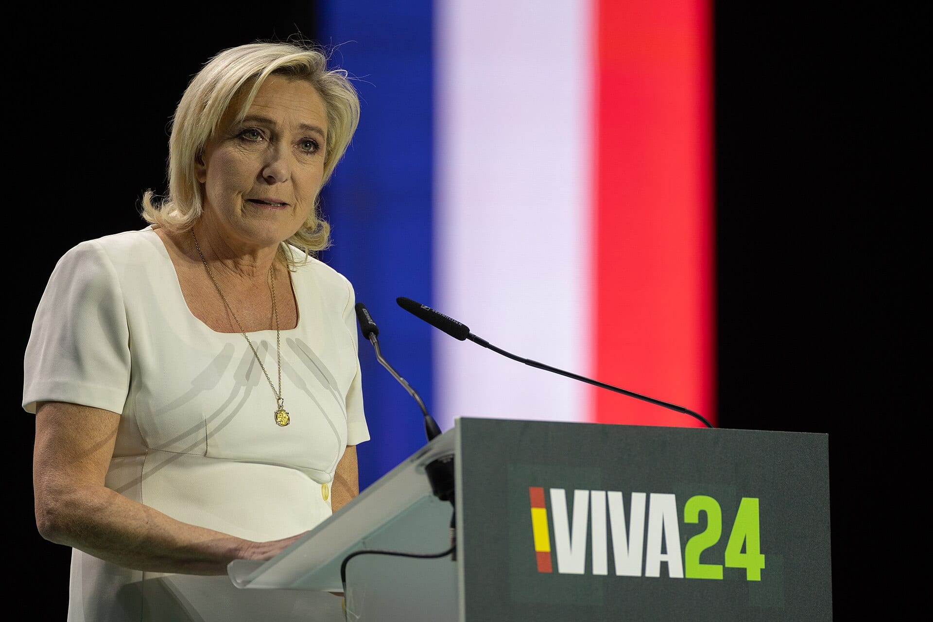 Marine Le Pen, de blanco, habla ante dos micrófonos en un atril, con una pantalla azul, blanca y roja detrás.