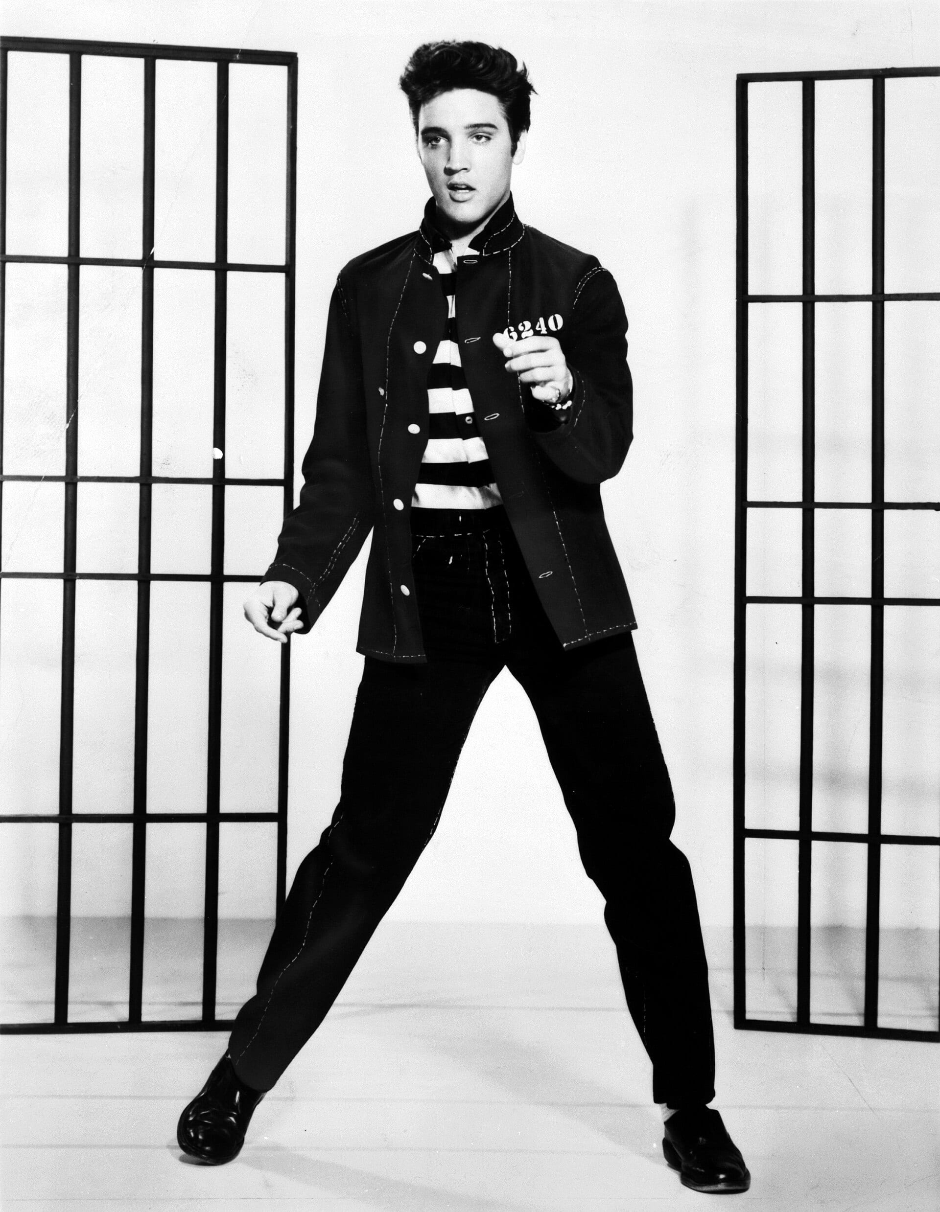 A black-and-white photo of Elvis Presley in a striped prison-style shirt and dark jacket, posing with his legs apart between two sets of bars.