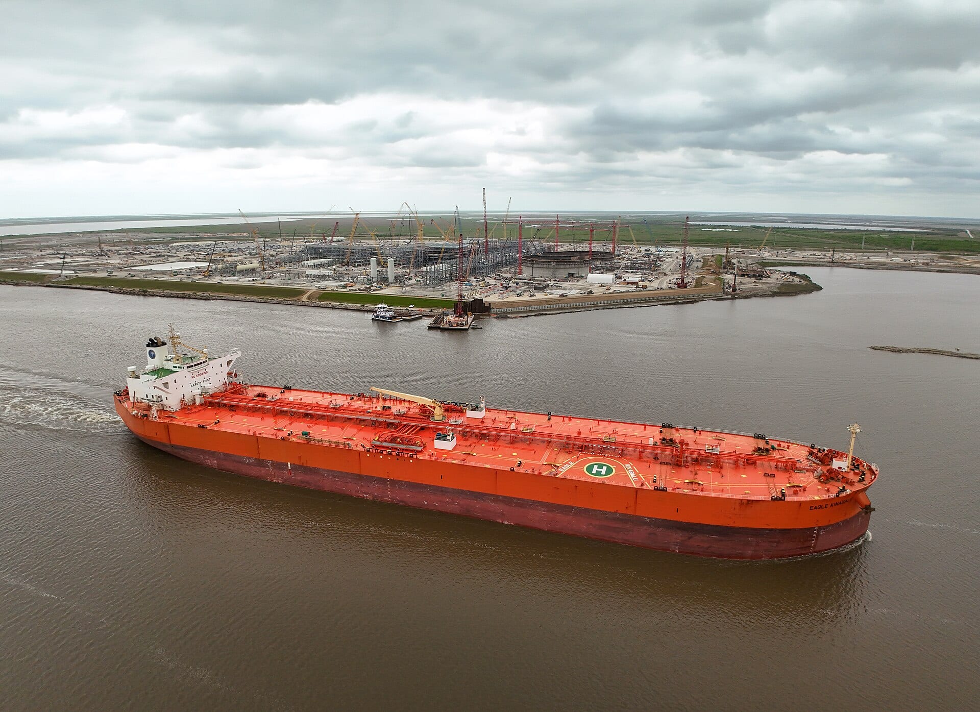 Un buque petrolero de casco naranja navega por un canal frente a una planta en construcción en la costa de Texas.