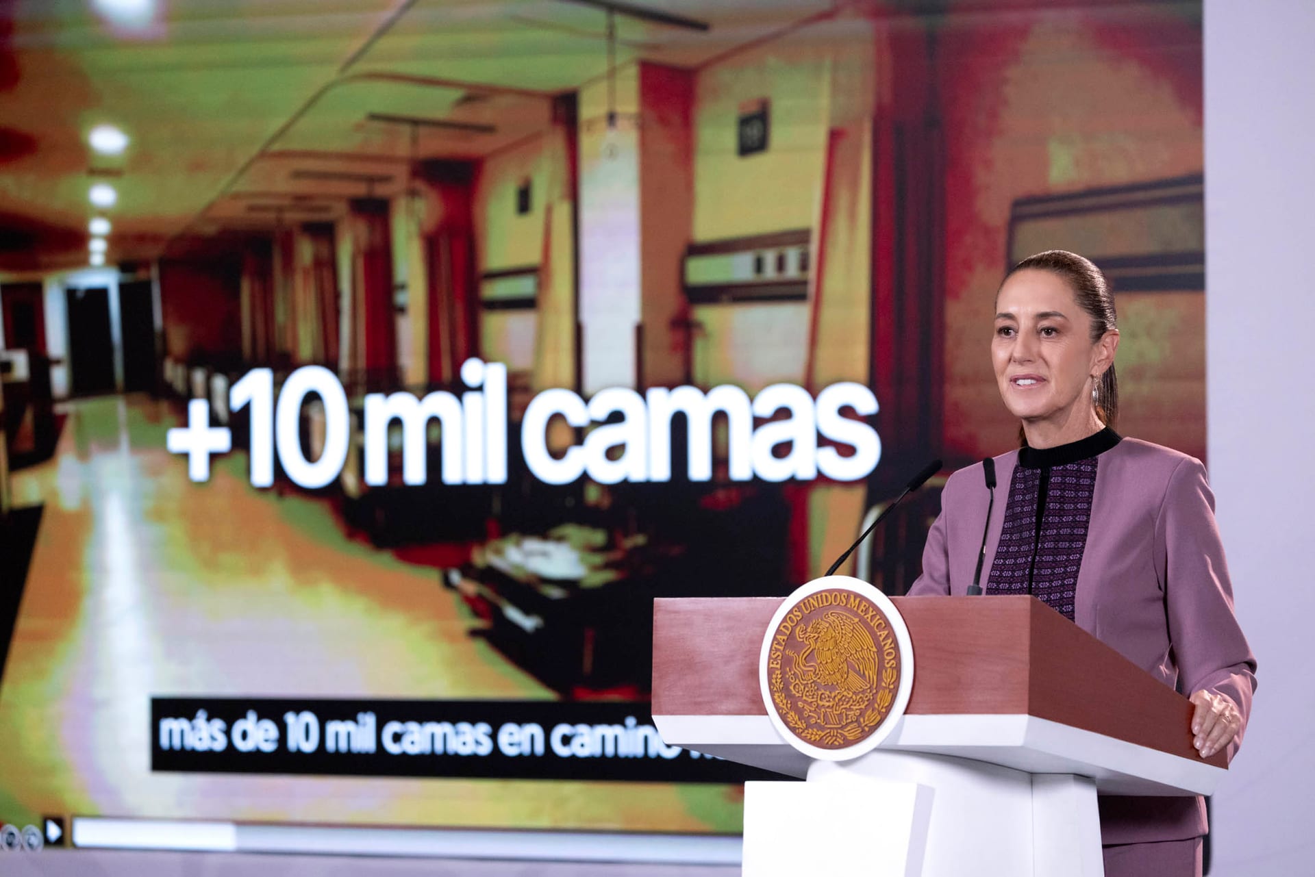 La presidenta Claudia Sheinbaum habla tras un podio con el escudo nacional mientras una pantalla detrás de ella muestra la leyenda más de 10 mil camas.