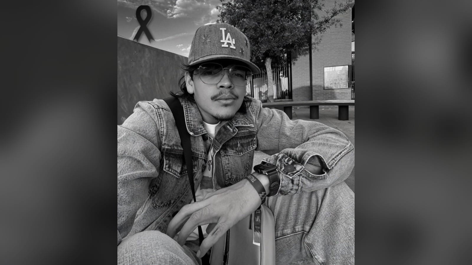 Hombre con gorra de los Dodgers, lentes de armazón metálico y chamarra de mezclilla, sentado al aire libre, en fotografía en blanco y negro, con un moño negro de luto en la esquina superior.