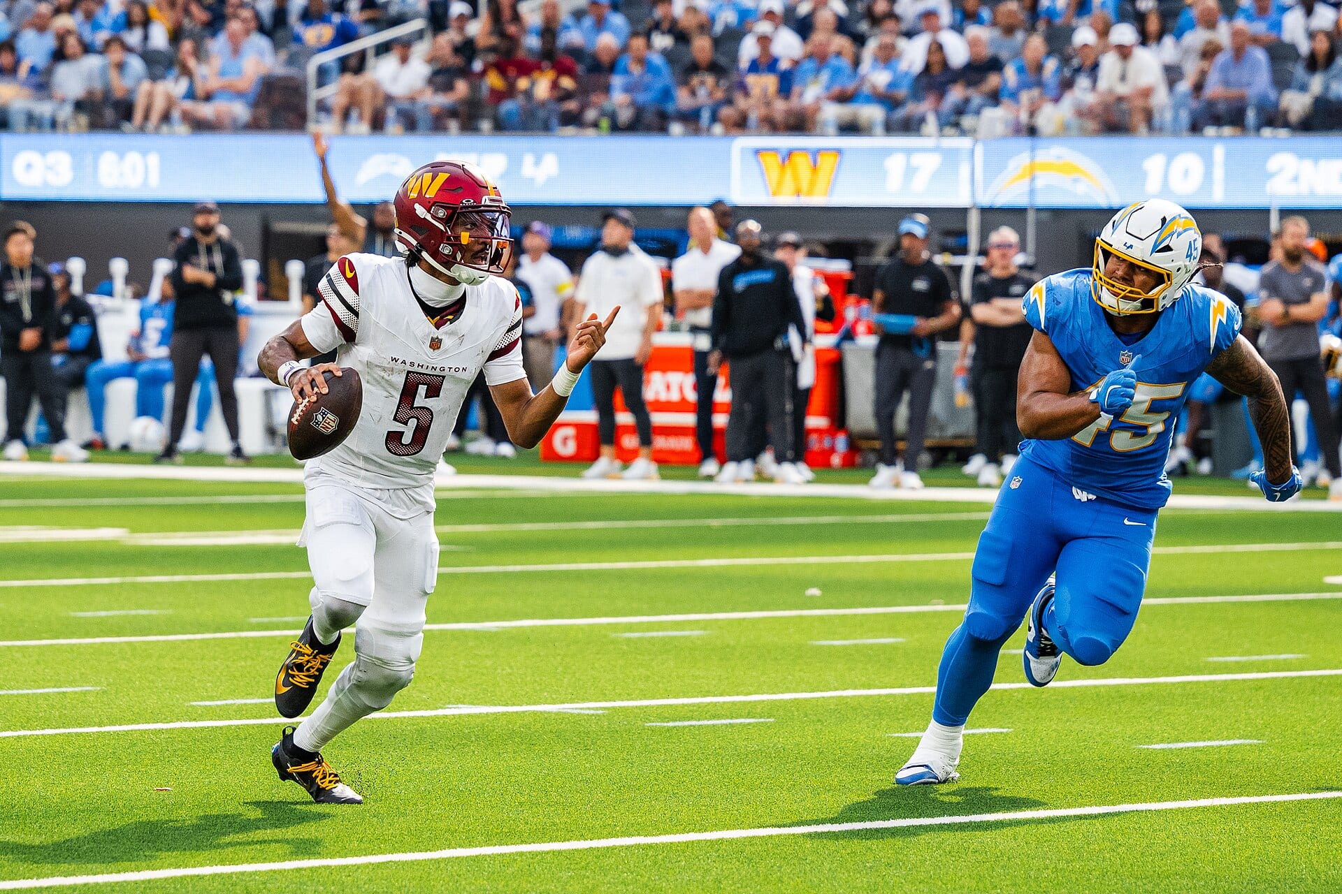 Washington's quarterback, in a white jersey with the number 5, scrambles out of the pocket with the ball in his hand while a defender in blue chases him.