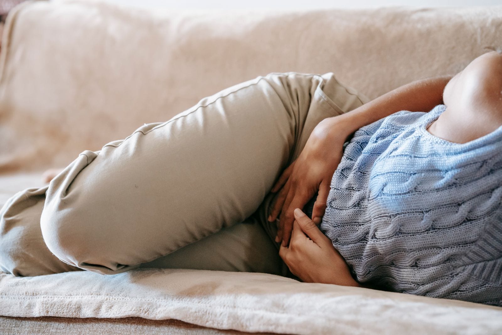 Una persona recostada de lado en un sofá claro, con suéter azul y las manos sobre el abdomen, sin mostrar el rostro.