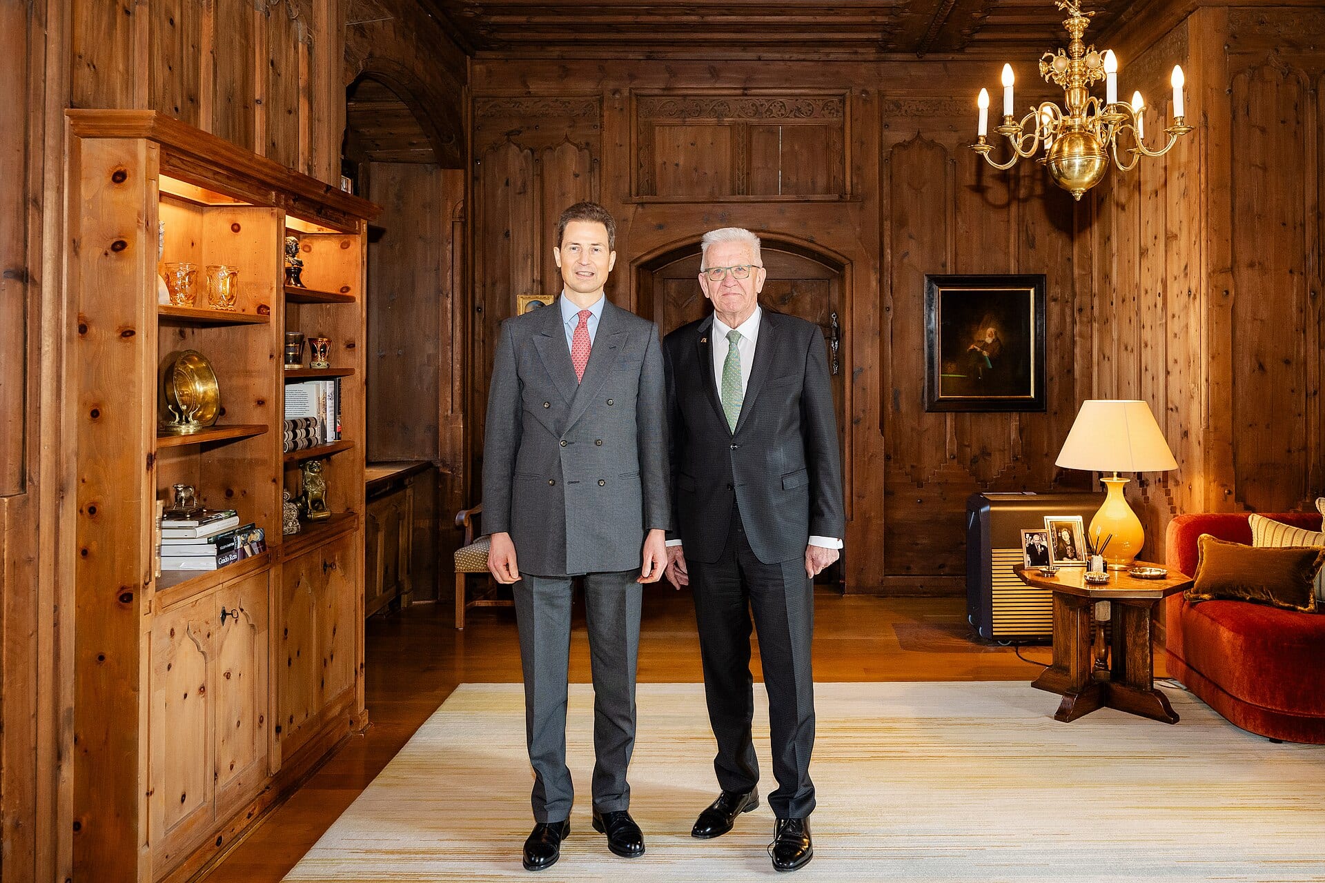 El príncipe heredero Alois, de traje gris, posa de pie junto a otro hombre en un salón con paredes de madera y una lámpara de bronce.