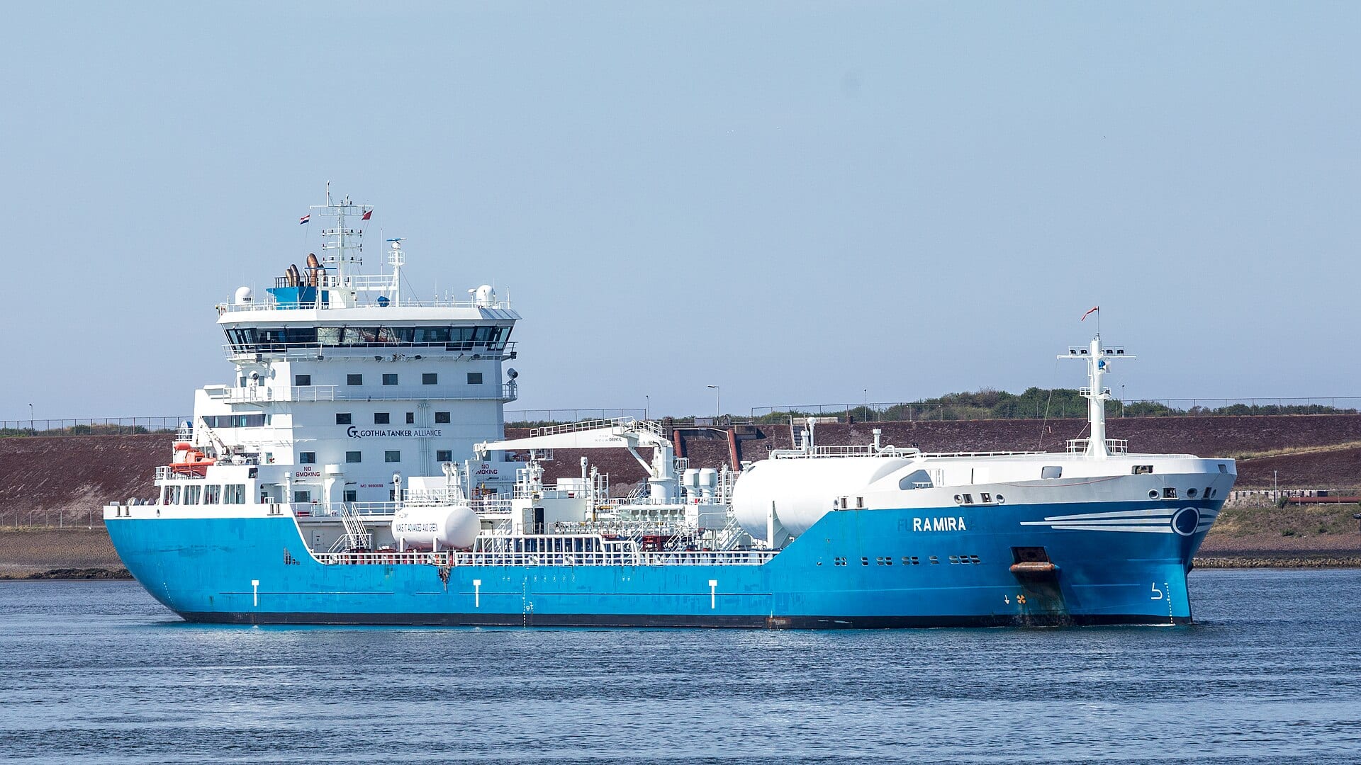 Un buque tanque de casco azul y superestructura blanca navega en aguas tranquilas frente a un dique.