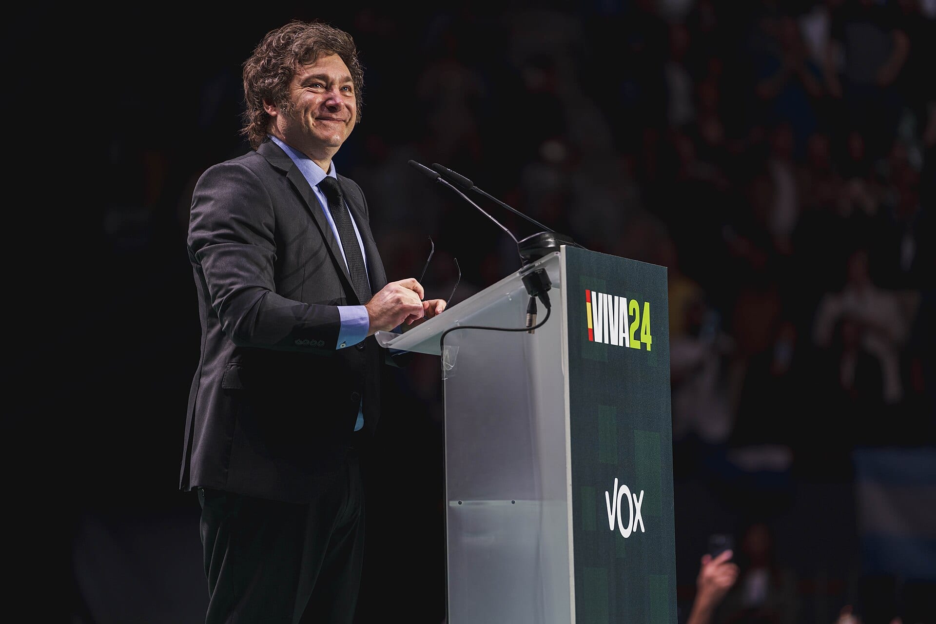 Javier Milei, in a dark suit, smiles at a clear lectern with two microphones while the crowd blurs in the background.