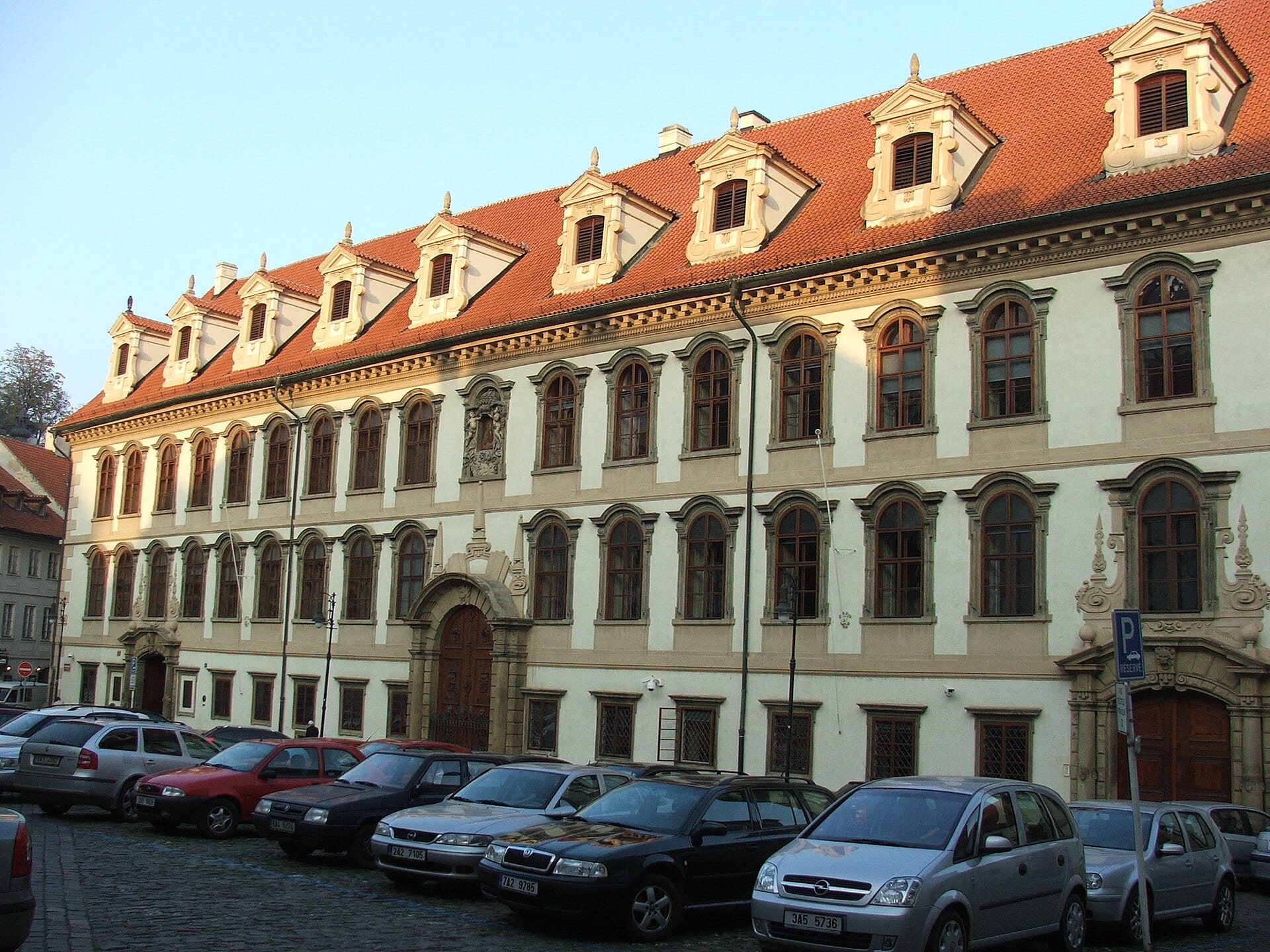 Facade of Wallenstein Palace, seat of the Czech Senate in Prague, with a red tile roof and parked cars in front.