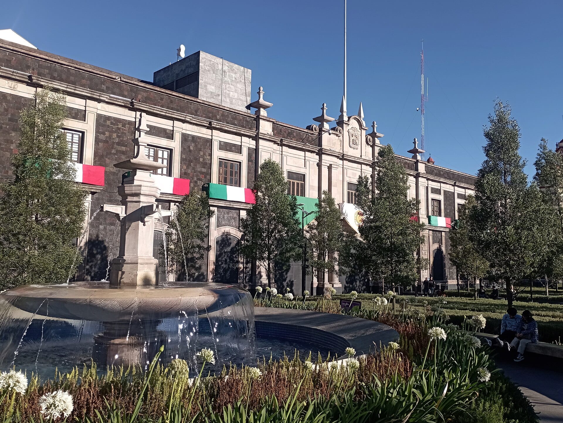 Fachada de piedra oscura del Congreso del Estado de México, con banderas tricolores en los balcones y una fuente en primer plano.