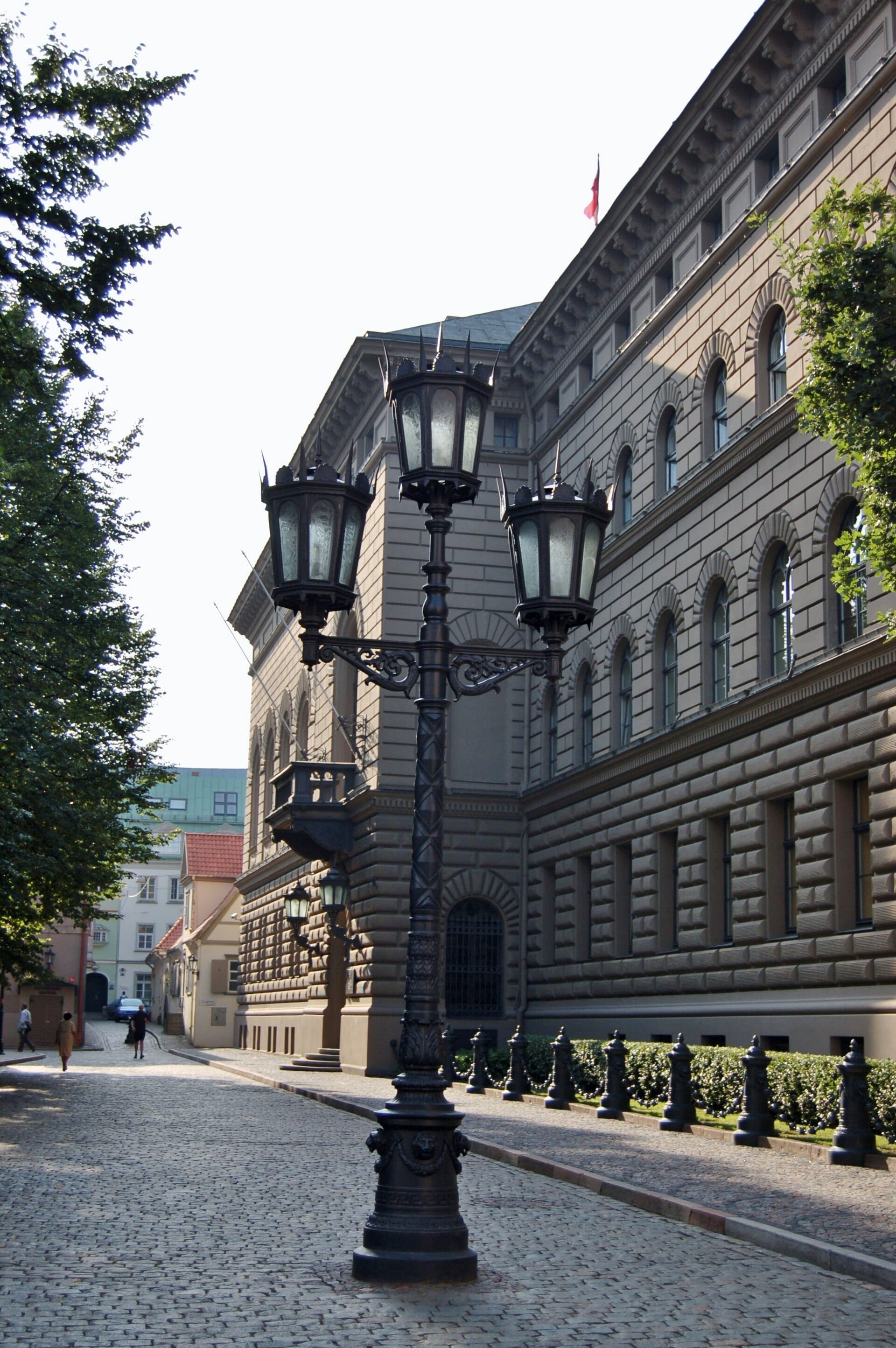 A wrought-iron lamp post with three lanterns in front of the stone Saeima building on a cobbled street in Riga.