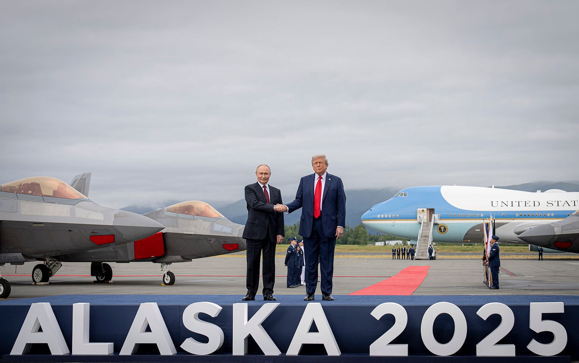 Vladimir Putin and Donald Trump shake hands on a stage, with fighter jets and the U.S. presidential plane behind them.
