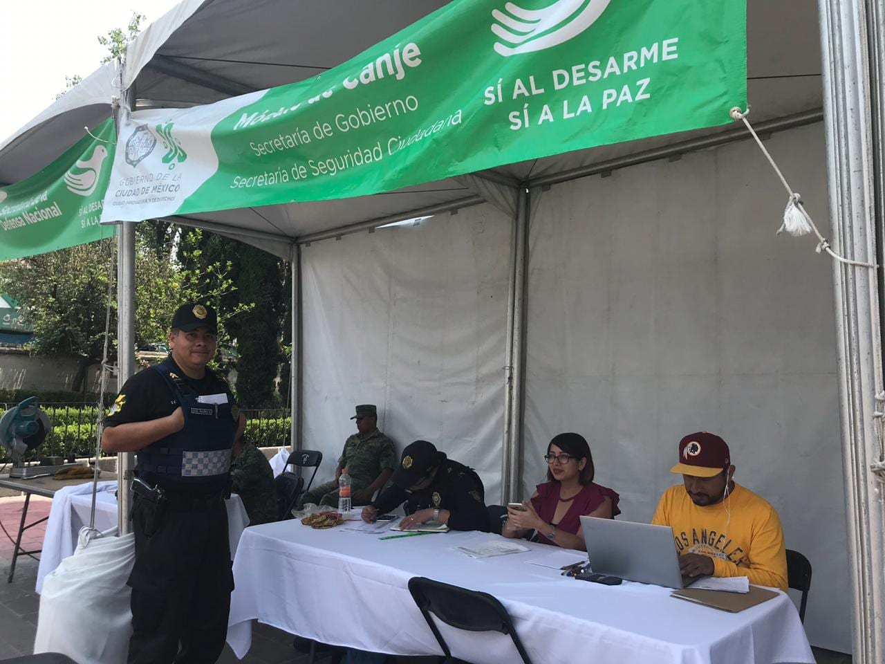 Carpa verde con el letrero de módulo de canje, "Sí al Desarme, Sí a la Paz", donde un policía de pie y personal sentado a una mesa registran datos.