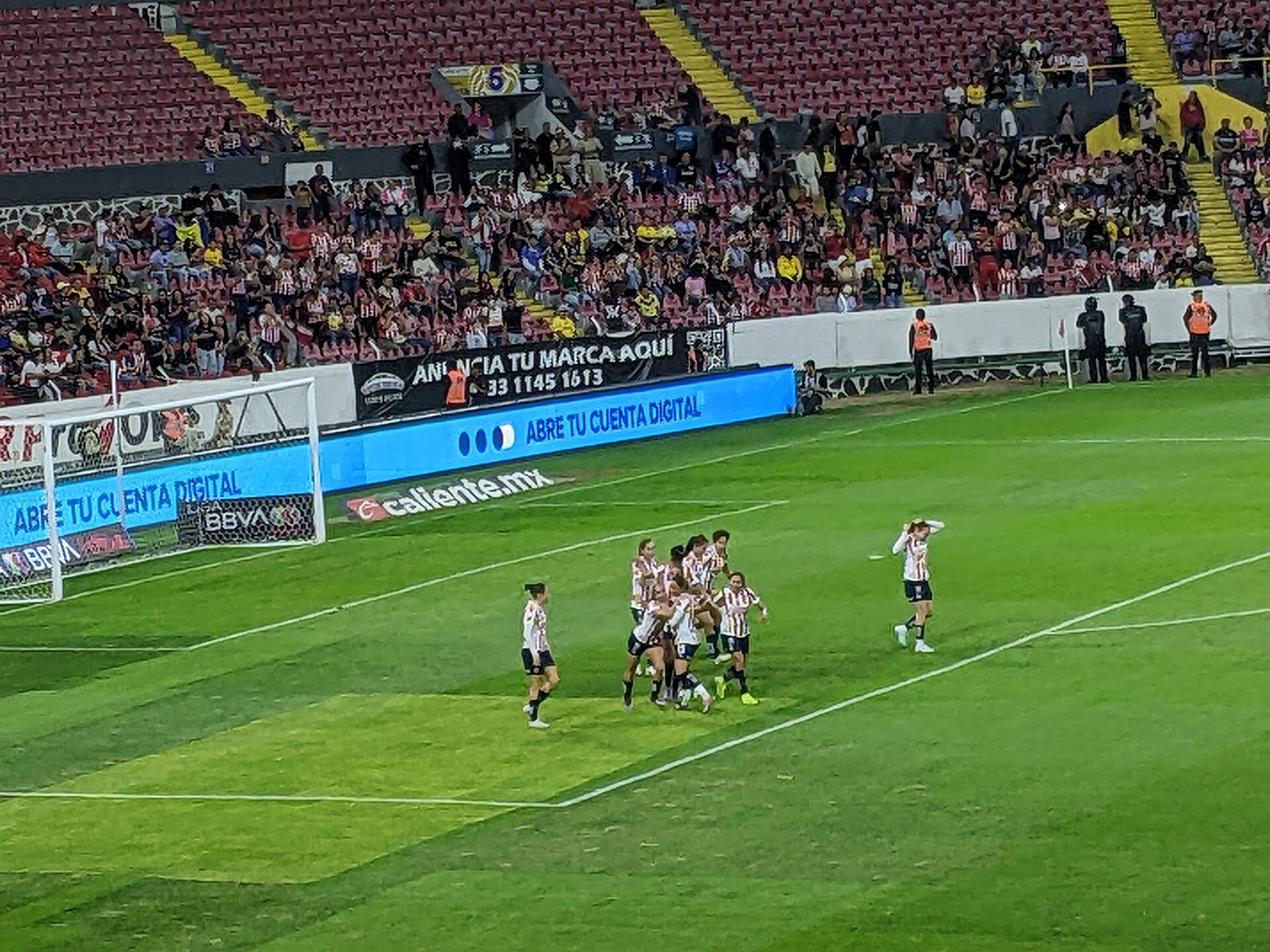 Jugadoras de Chivas con uniforme rojiblanco se abrazan en grupo tras anotar, frente a las gradas del estadio.