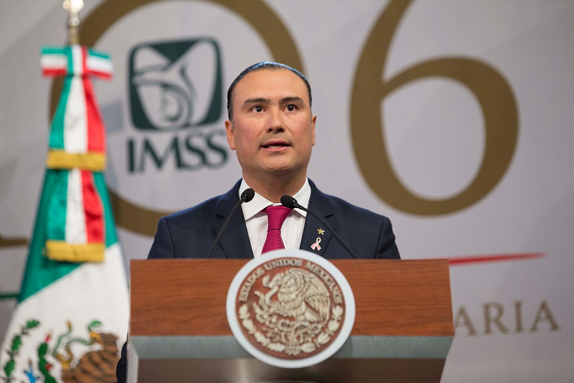 Un orador de traje oscuro habla desde un podio con el escudo nacional, frente a una bandera de México y el logotipo del IMSS.