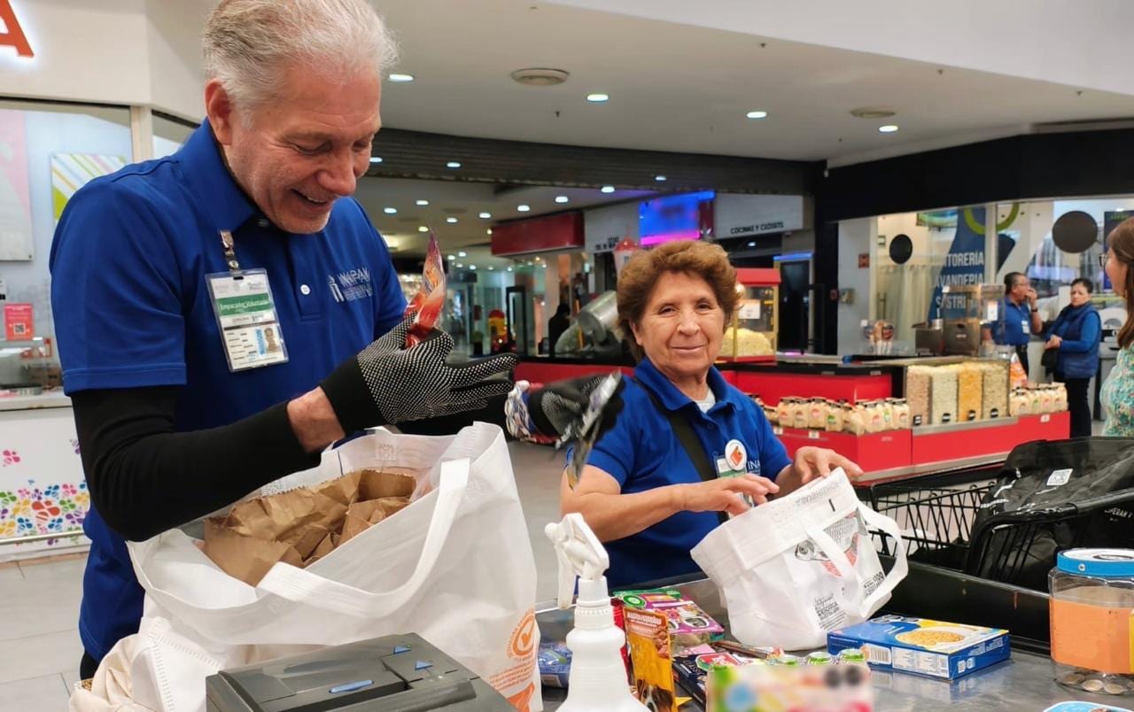 Dos personas mayores con playera azul empacan productos en bolsas blancas en la caja de una tienda.