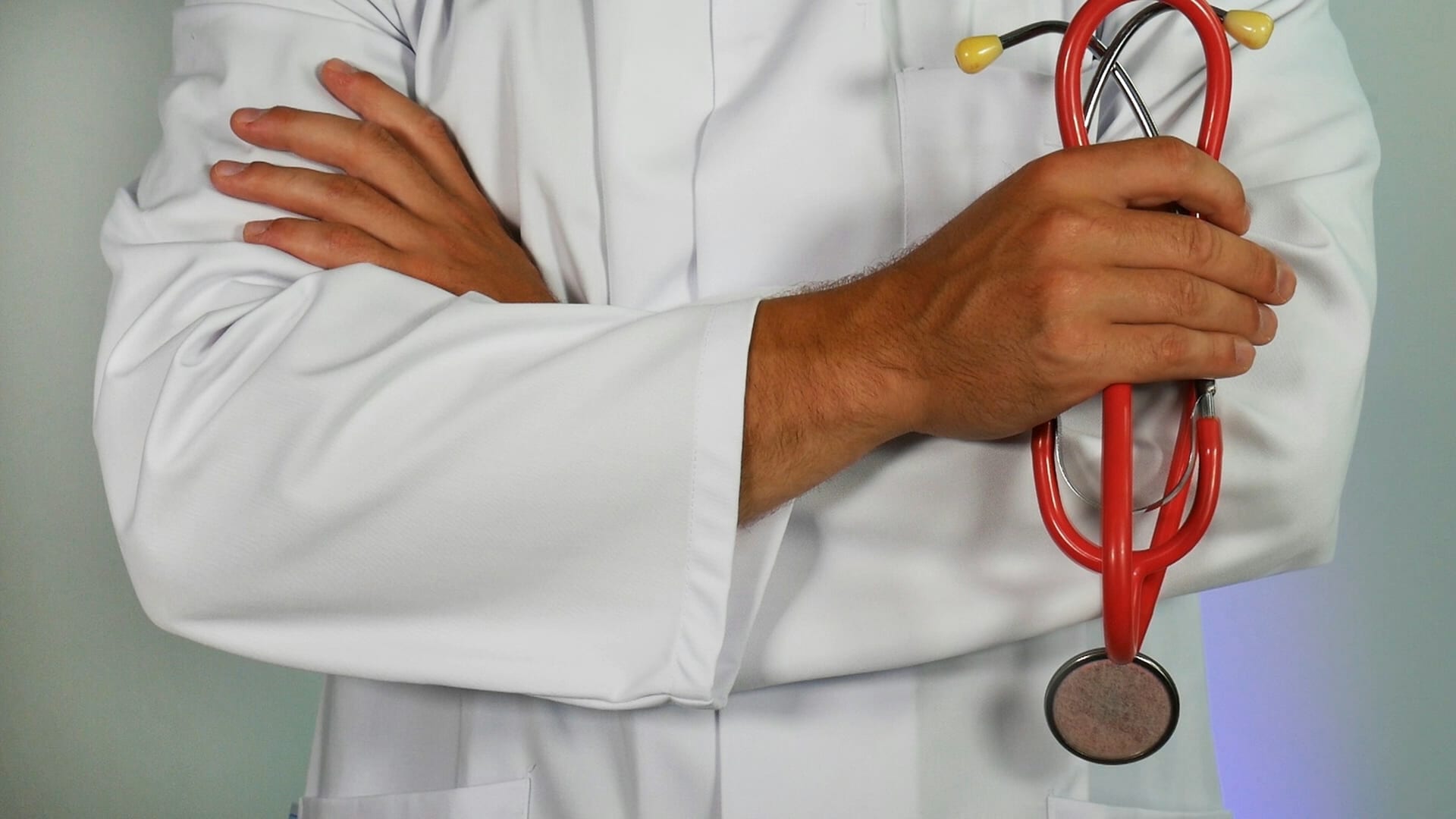 Un profesional de salud con bata blanca y los brazos cruzados sostiene un estetoscopio rojo.