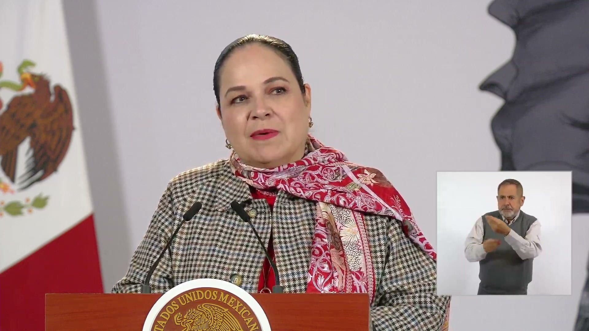 Una mujer de chaqueta de cuadros y bufanda roja habla detrás de un atril con el escudo nacional; a la derecha, un intérprete de lengua de señas.