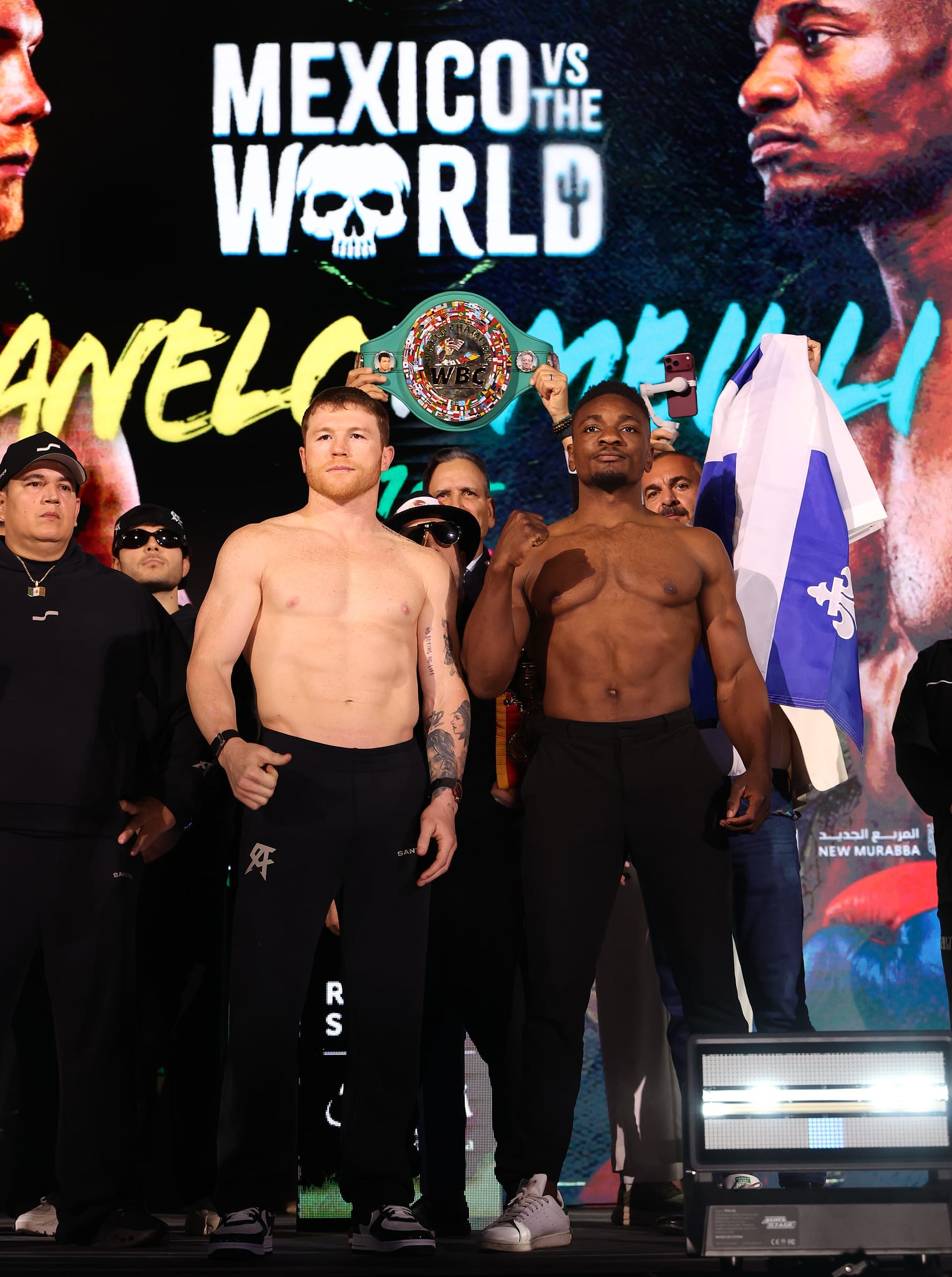 Canelo Alvarez and Christian Mbilli pose shirtless at the Riyadh weigh-in, with the green WBC belt above them and a screen with their names behind.