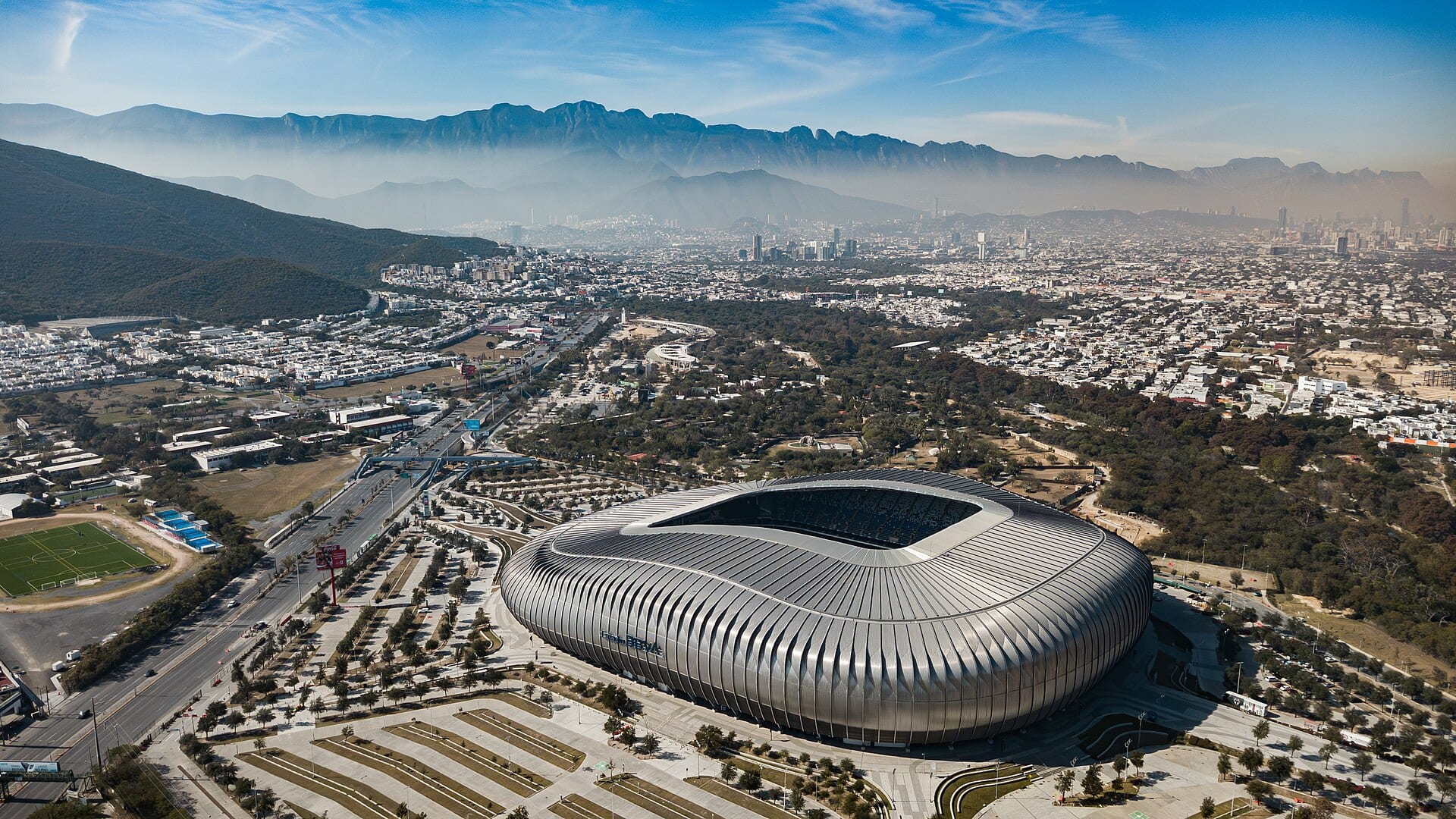 Vista aérea del Estadio BBVA, de fachada metálica ondulada, con la ciudad de Monterrey y las montañas al fondo.