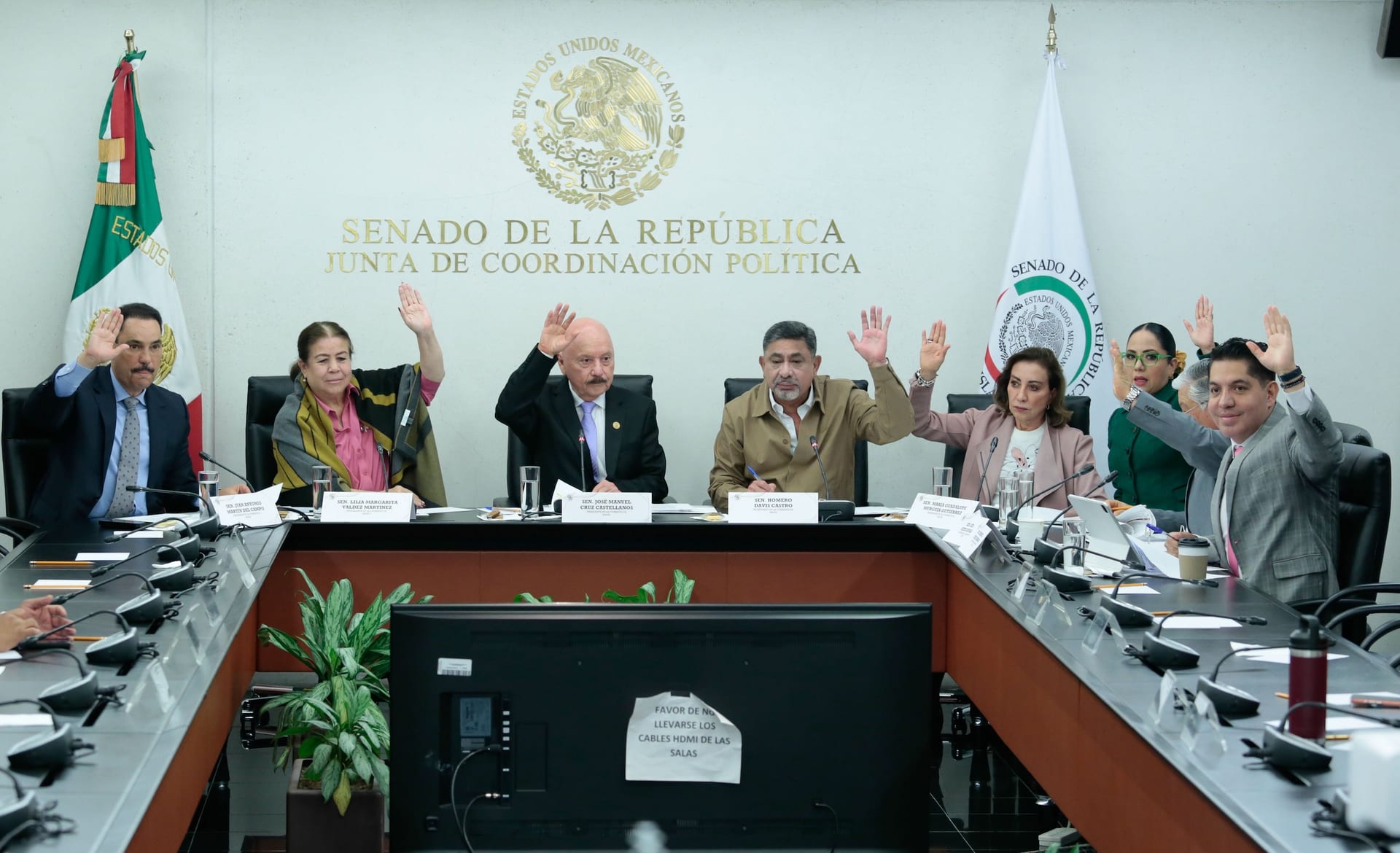 Senadoras y senadores sentados a una mesa en forma de U levantan la mano para votar, frente al escudo y el letrero del Senado.