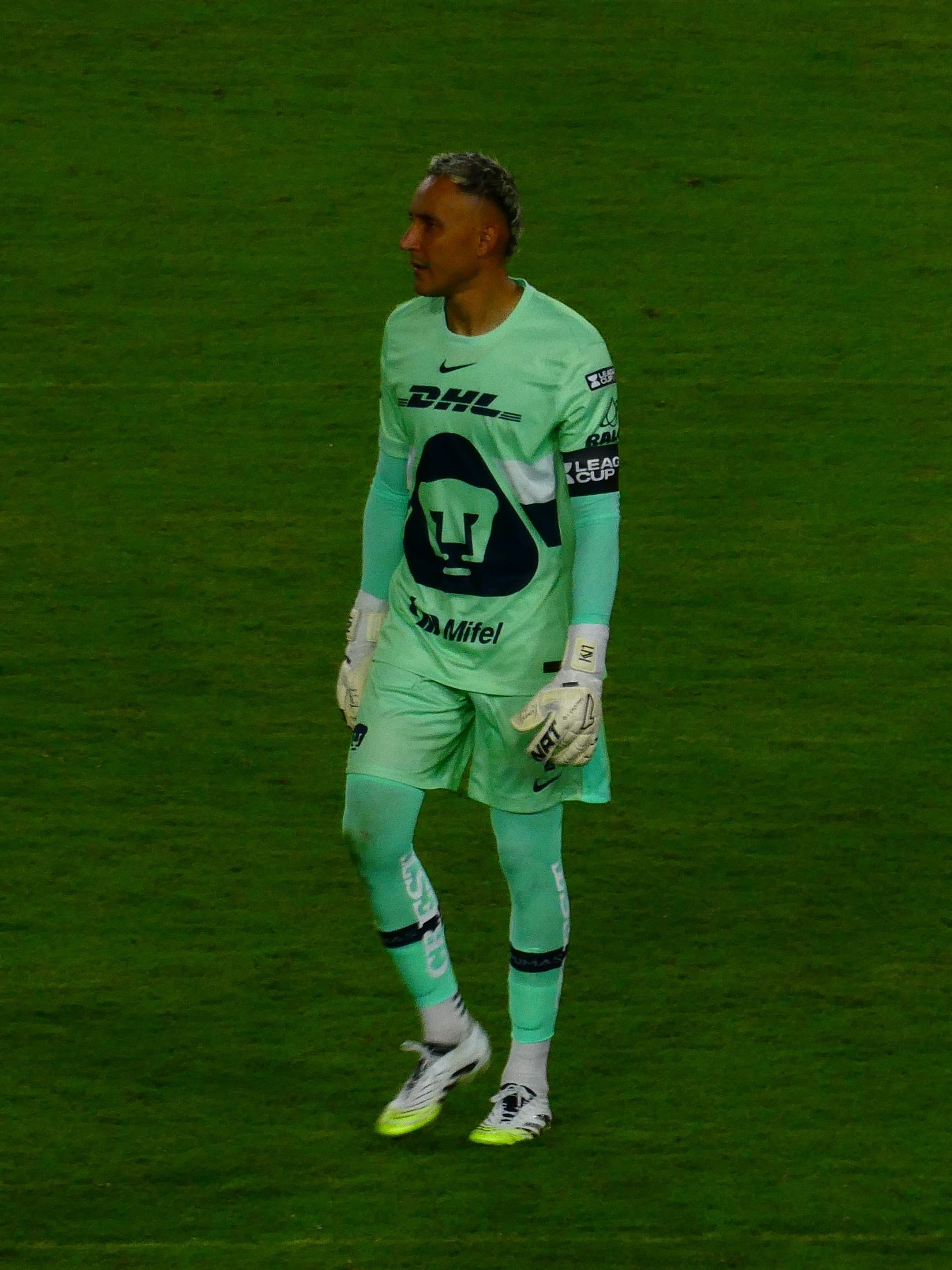 Keylor Navas, con uniforme verde claro y guantes, camina solo sobre el césped durante un partido de Pumas.