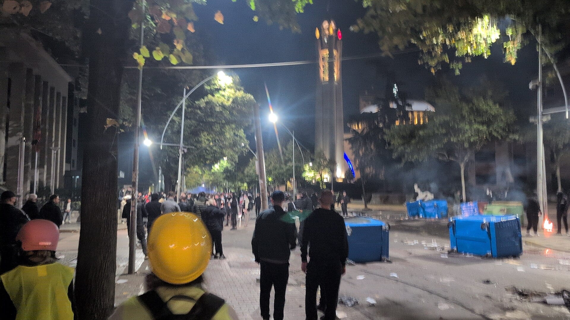 Una calle de Tirana de noche, con manifestantes al fondo, contenedores azules volcados sobre el pavimento y una torre iluminada.