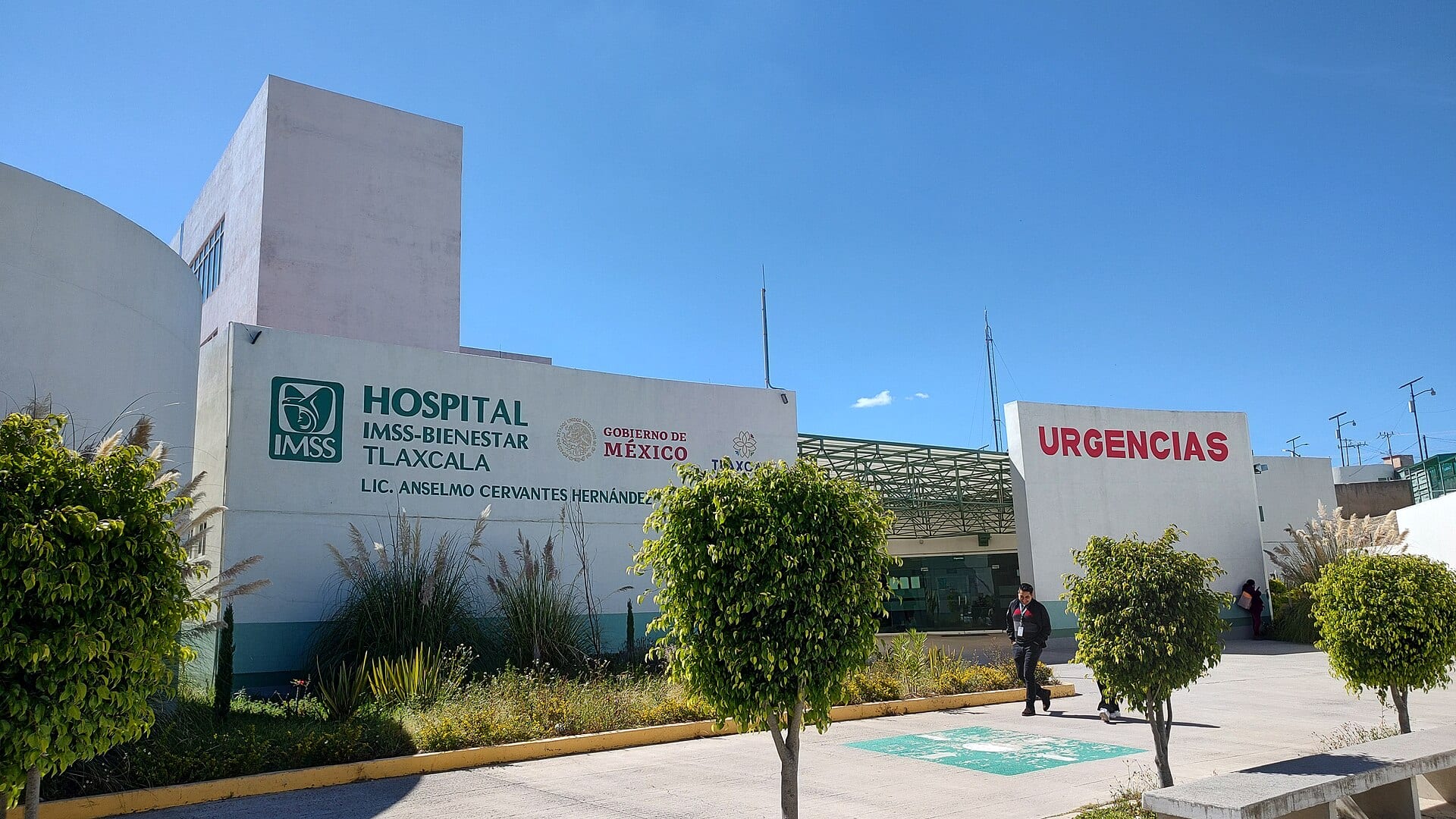 Fachada blanca de un hospital con el rótulo IMSS-Bienestar Tlaxcala y, a la derecha, la entrada de Urgencias, entre arbustos podados.