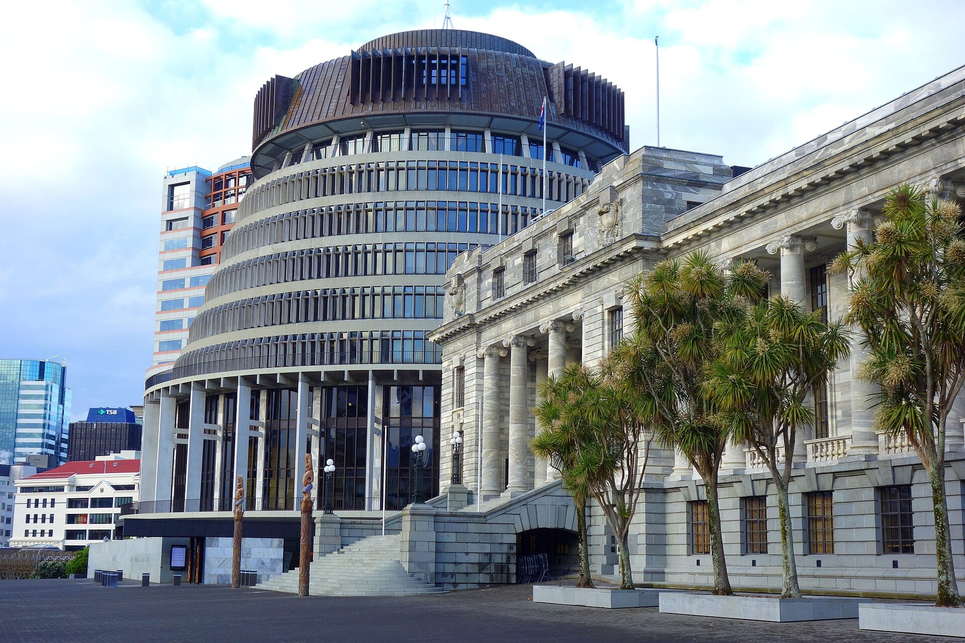 El edificio circular del Beehive y la antigua sede del Parlamento en Wellington, con palmeras al frente y una plaza empedrada en primer plano.