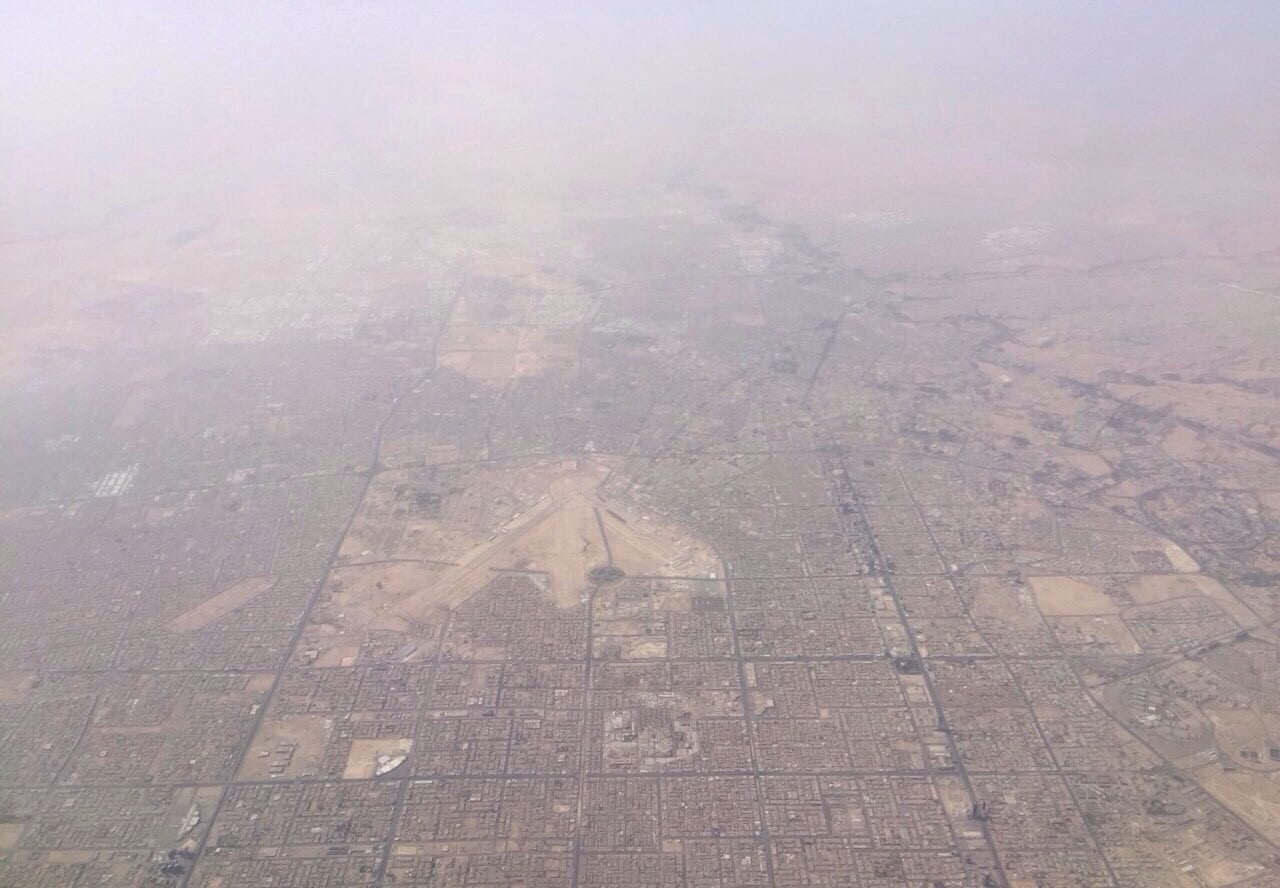 Vista aérea entre la calima de una ciudad de calles en cuadrícula en el desierto, con una explanada de pistas al centro.