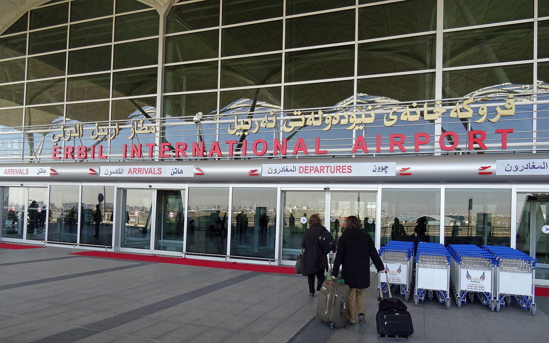 Fachada de la terminal del aeropuerto internacional de Erbil, con dos pasajeros que arrastran maletas hacia la entrada.