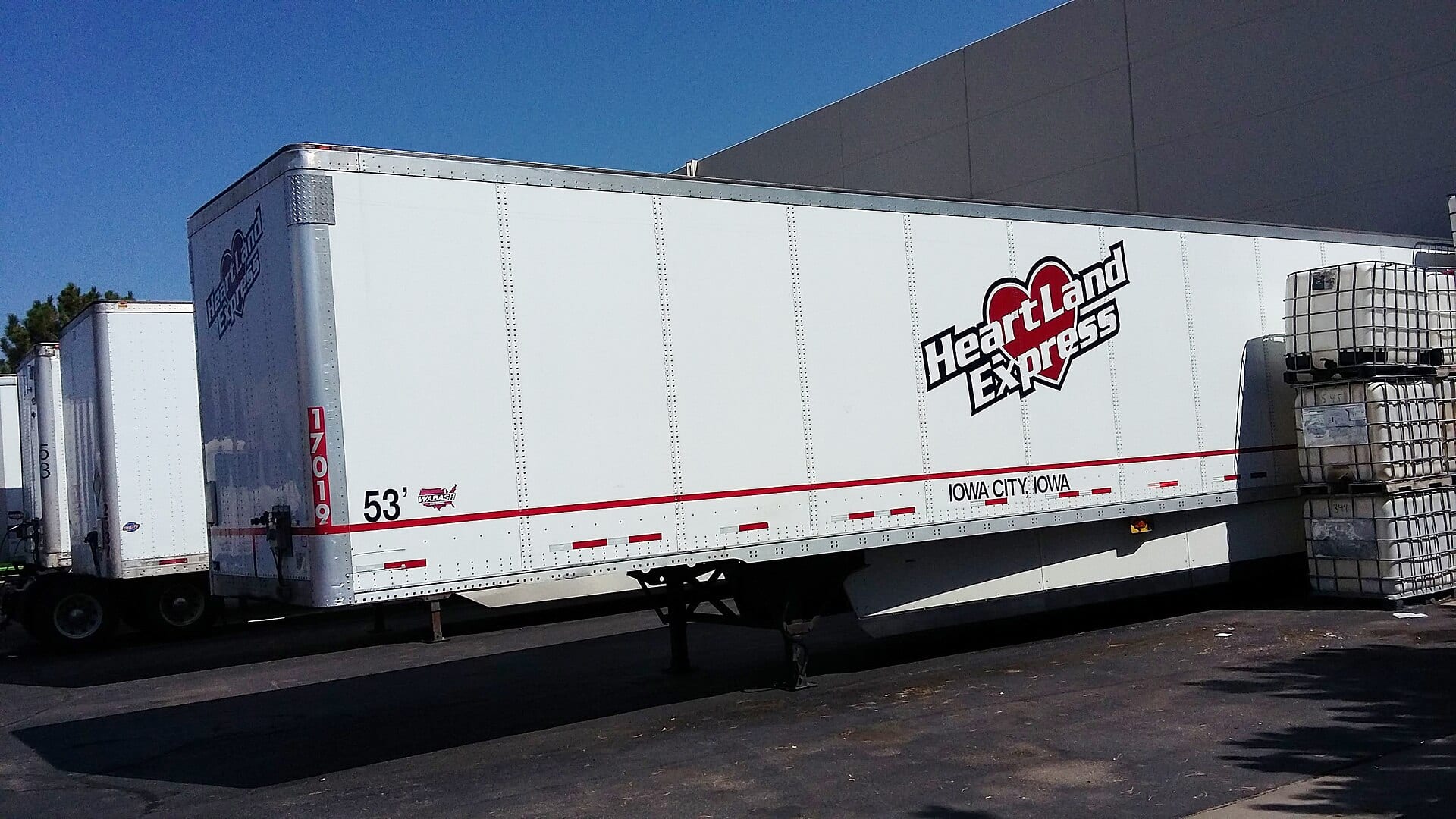 A white 53-foot van trailer parked beside a warehouse, with more trailers behind it.