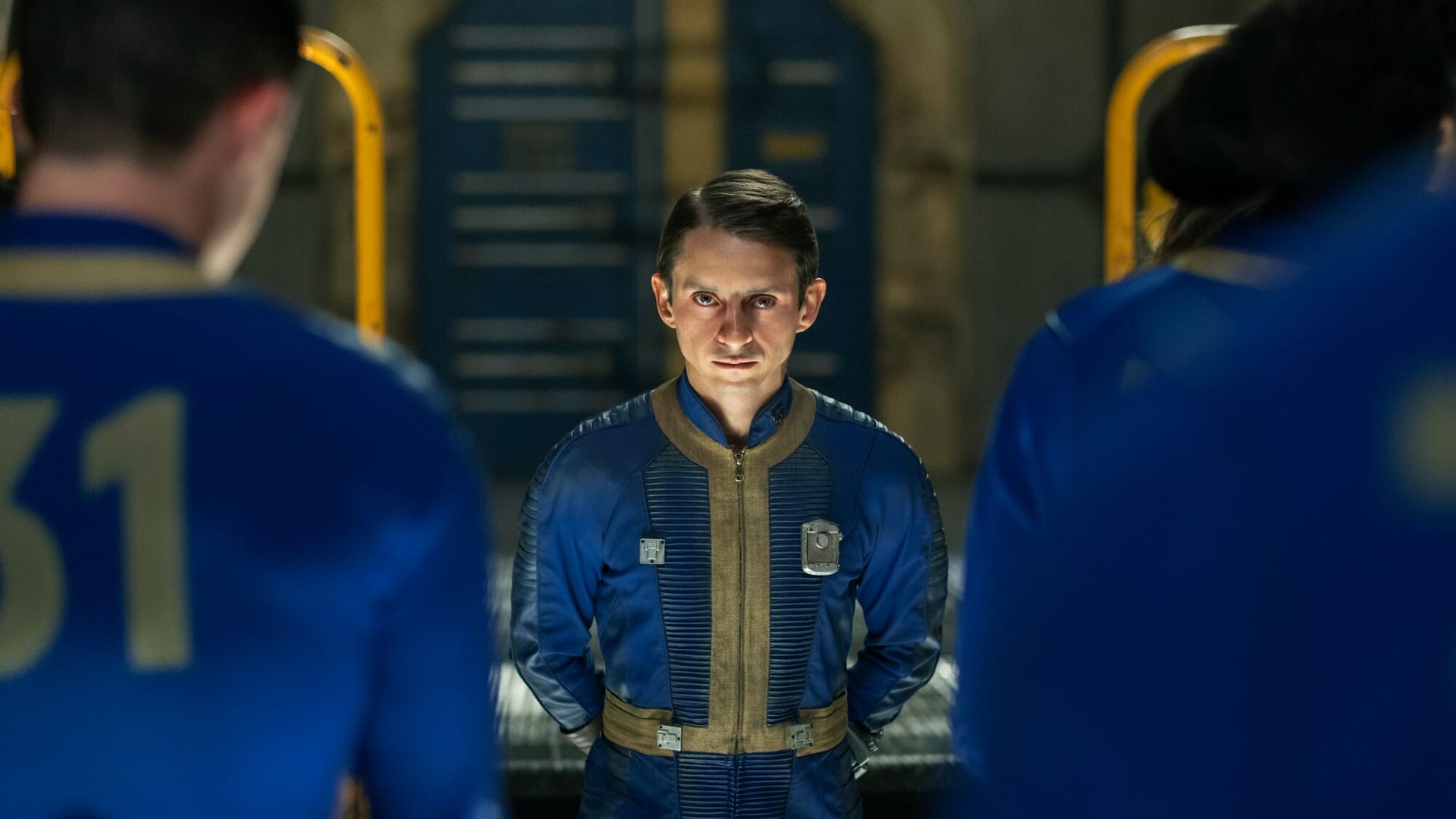Un hombre con el traje azul y dorado de Vault-Tec mira a cámara entre dos figuras desenfocadas, imagen de la serie Fallout de Prime Video.