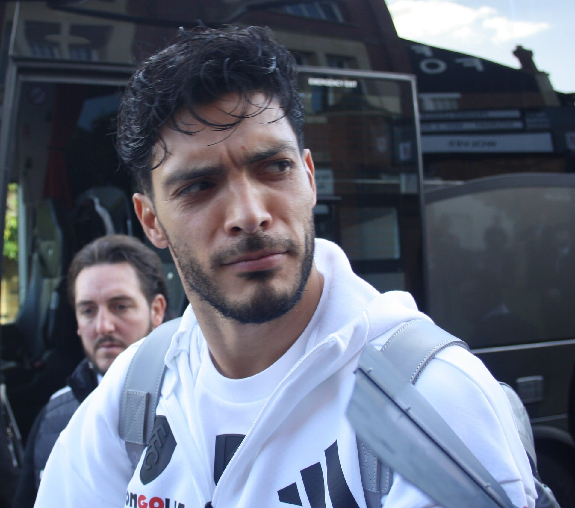 Raúl Jiménez, de cabello oscuro y barba, mira hacia un lado junto a un autobús; lleva una sudadera blanca y una mochila gris.