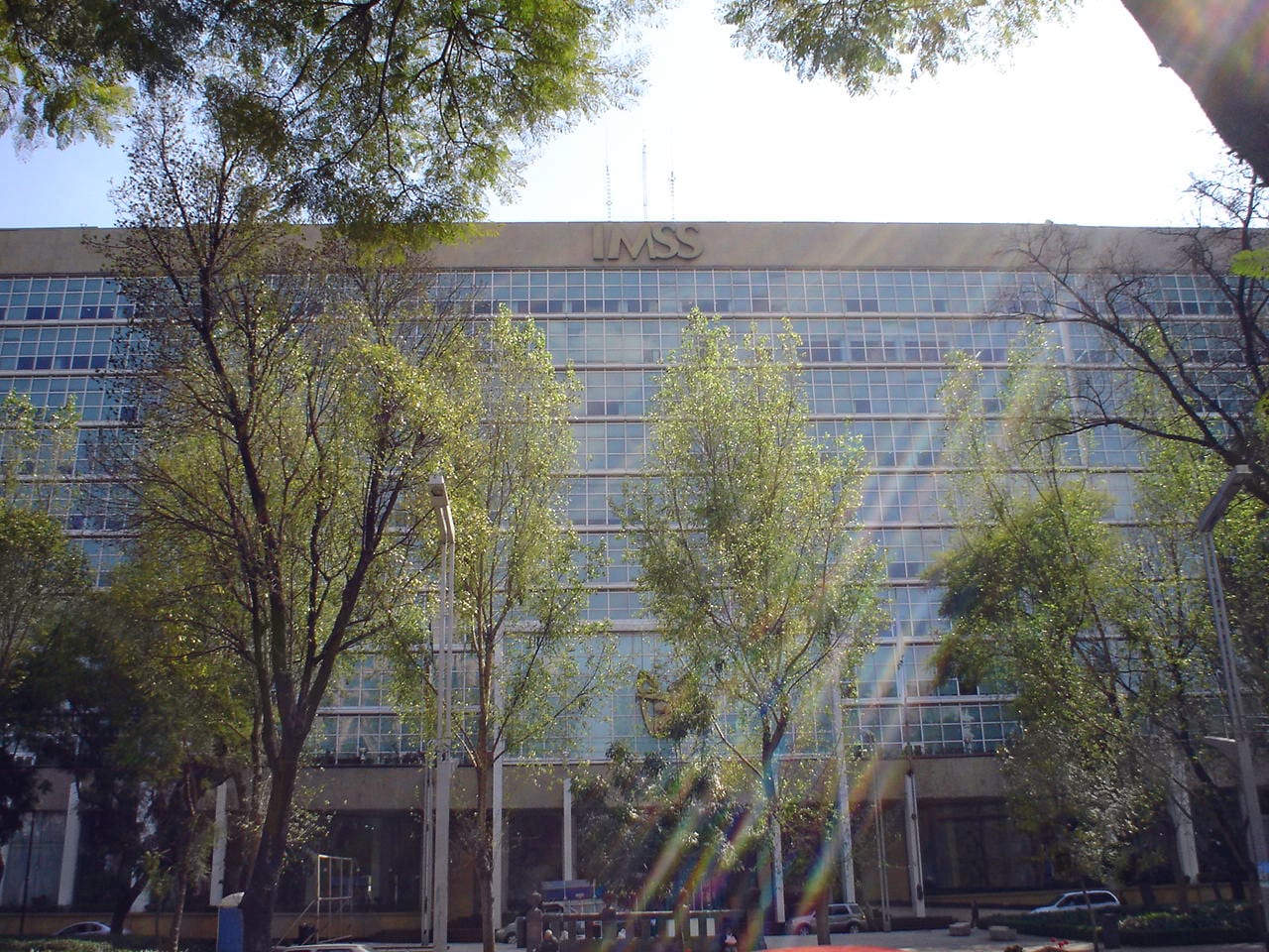Fachada de vidrio del edificio de oficinas centrales del IMSS con las letras IMSS en lo alto, vista entre árboles.