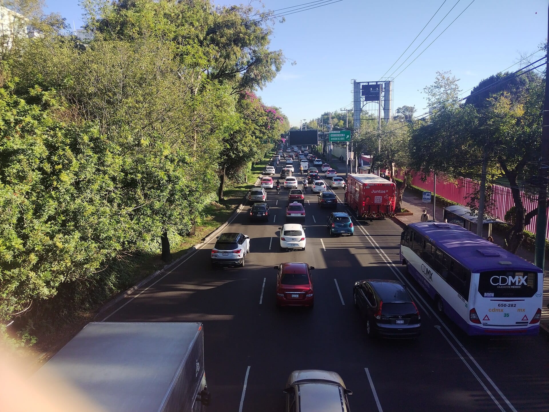Una avenida de varios carriles con tráfico, un autobús de la ciudad y árboles a los lados, vista desde un puente peatonal.