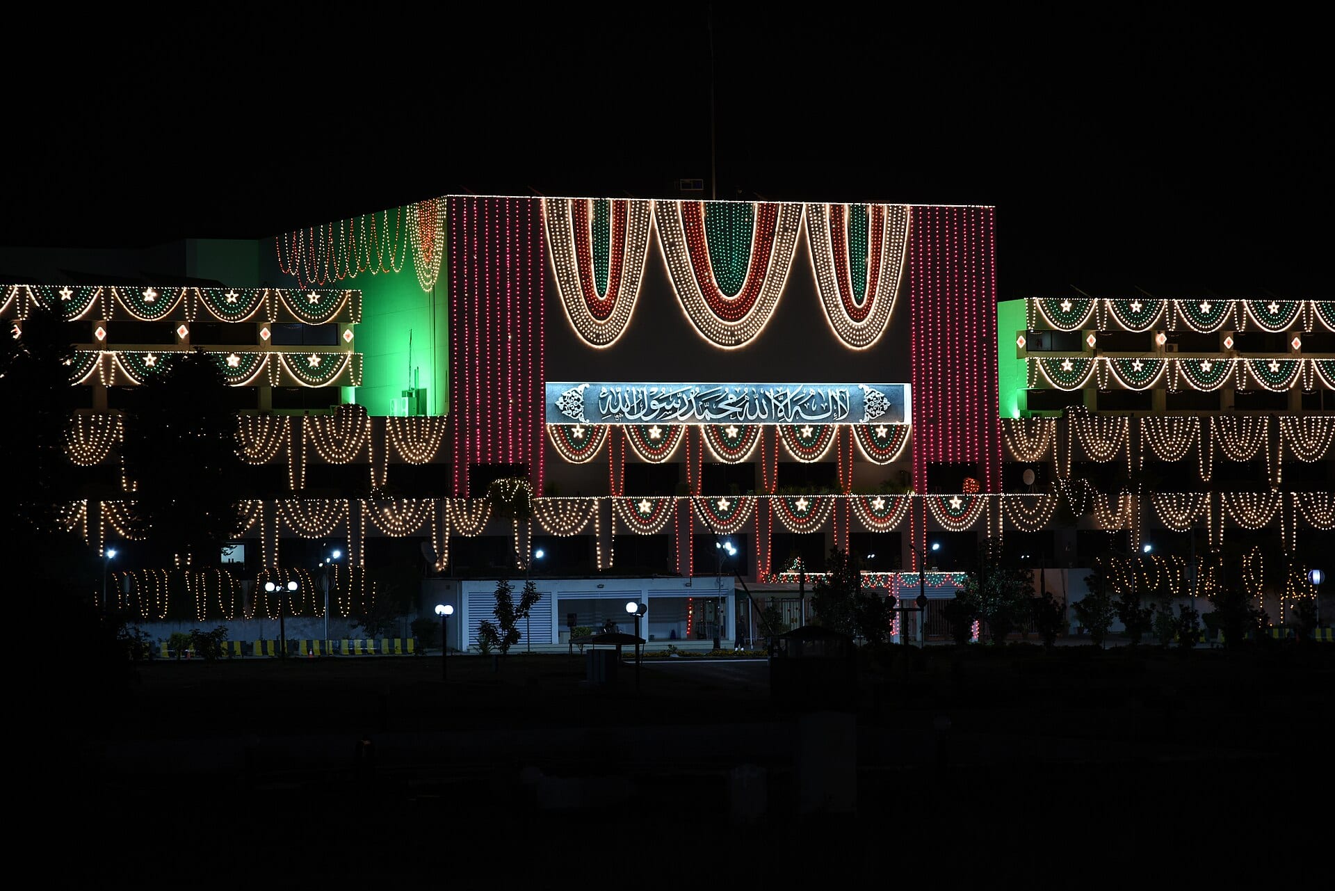 Night view of the Parliament building in Islamabad, covered in strings of green, red and white lights, with a panel of Arabic calligraphy at the center.