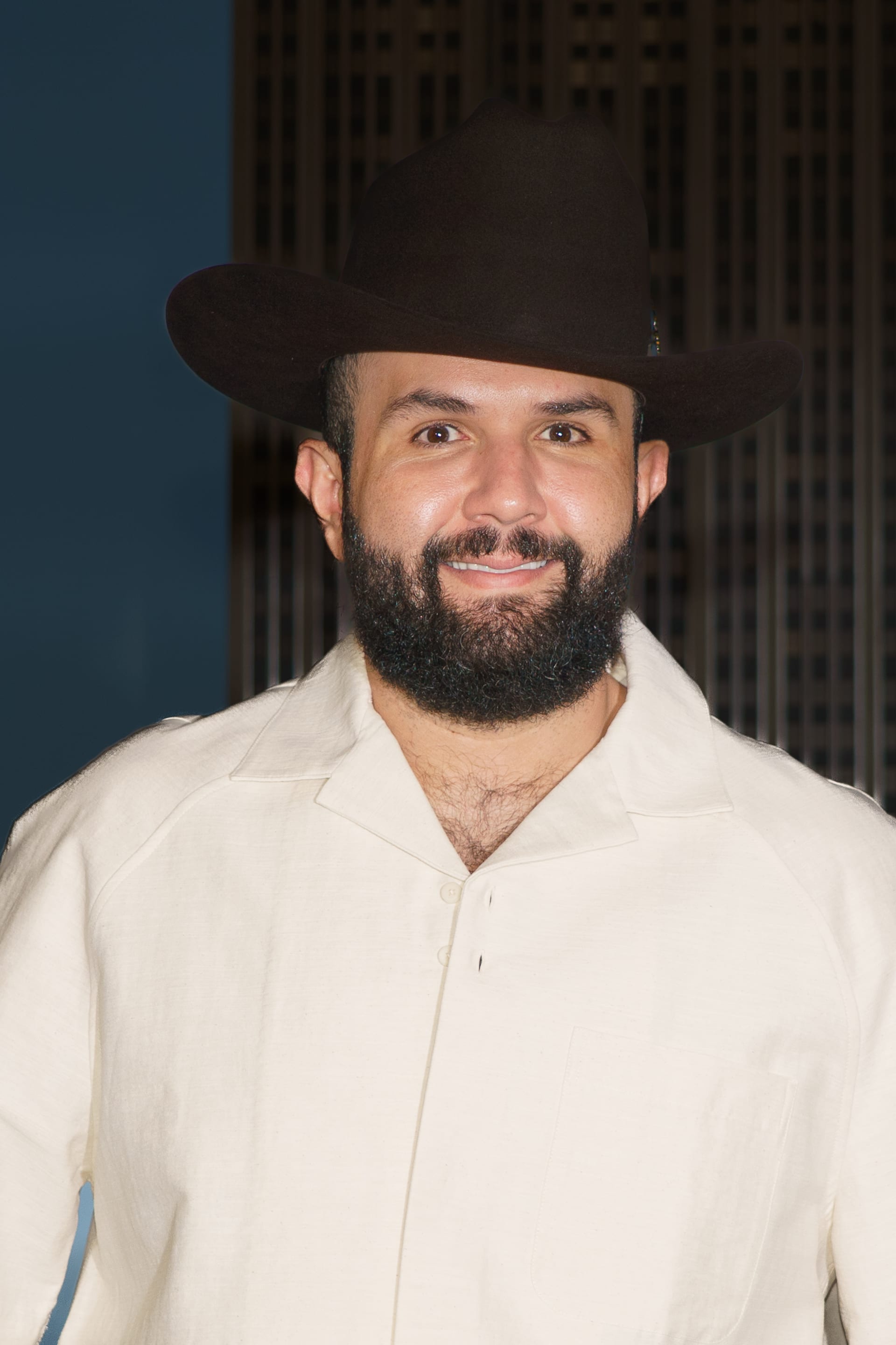 Carín León, con sombrero vaquero oscuro y camisa blanca, sonríe frente a un fondo azul y oscuro.