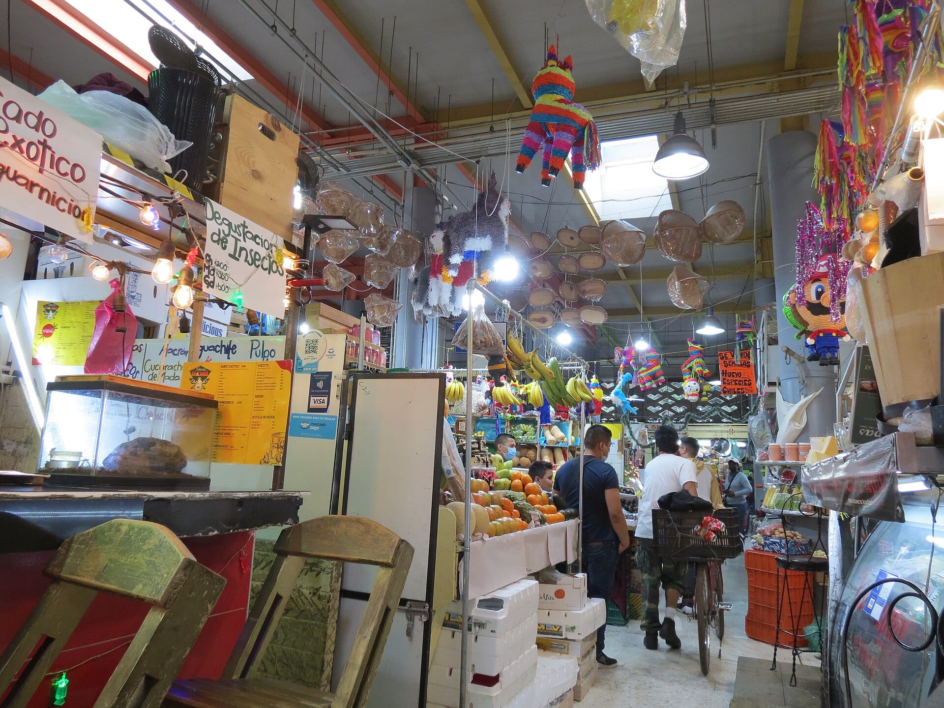 Pasillo del Mercado de San Juan con puestos de fruta, piñatas colgadas del techo y varias personas caminando entre los locales.