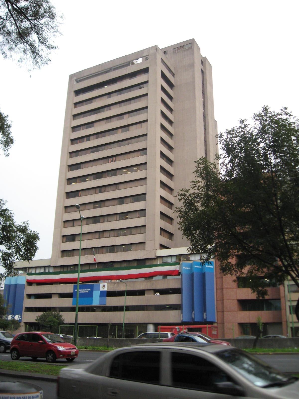 Torre de concreto de unos quince pisos, con una manta tricolor y paneles azules de Profeco en la base, junto a una avenida.